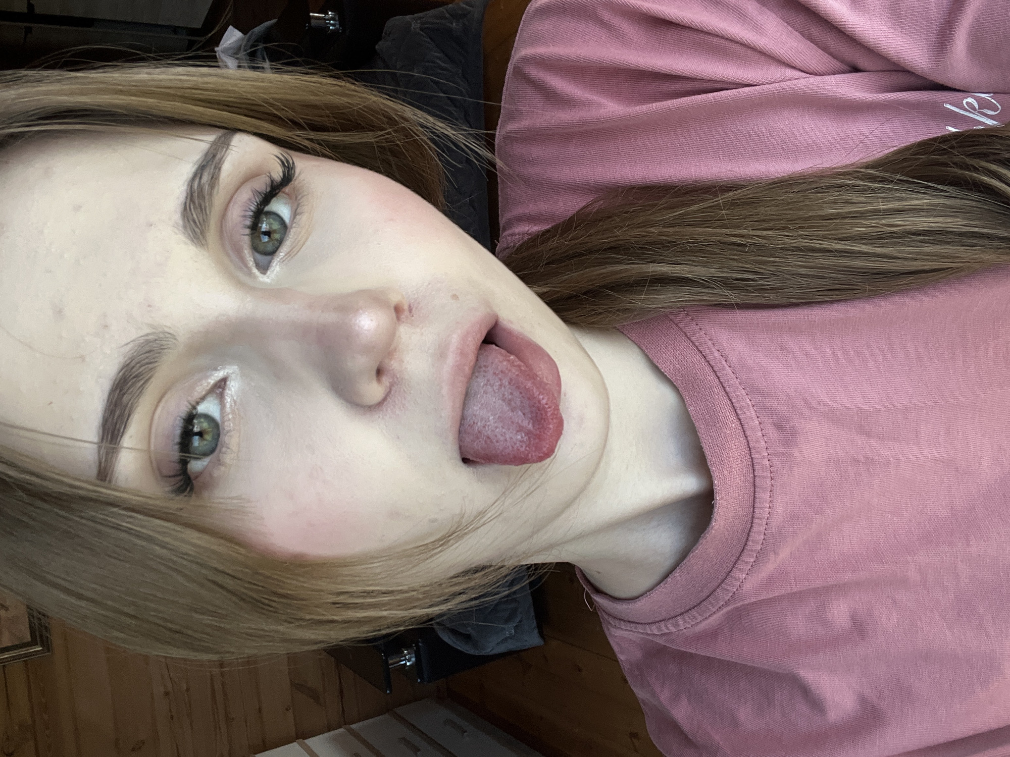 A close-up selfie of a young Caucasian woman with light brown hair and green eyes. She is wearing a pink t-shirt and has her tongue sticking out. Her makeup includes blush and mascara. The background appears to be an indoor setting with wooden walls and furniture, possibly a bedroom or living area. The image quality is moderate, with natural lighting. There is no indication of the specific location within the city of Ashkadar, Russia.