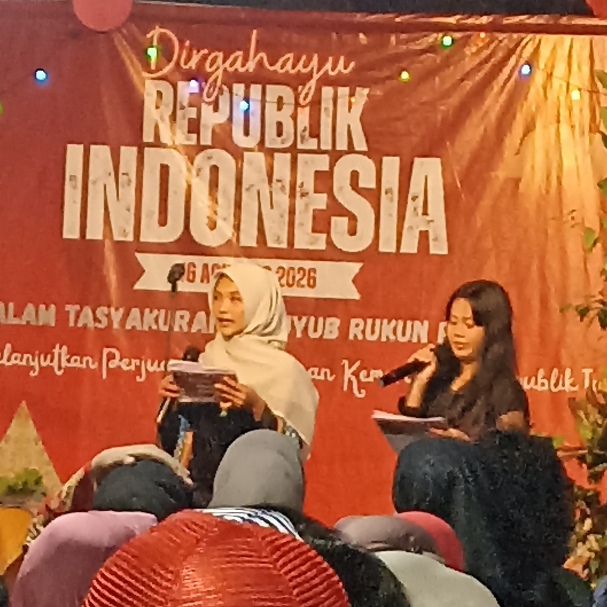 The image depicts a dimly lit indoor or outdoor evening event, possibly a celebration. A large orange banner with white text serves as the backdrop, reading "Dirgahayu REPUBLIK INDONESIA" and "G A 2026". Below this, smaller text is visible, including "ALAM TASYAKURA" and "lanjutkan Perju". Two women are the main subjects. The woman on the left, wearing a light-colored hijab, is holding and reading from documents while speaking into a microphone. The woman on the right, with long dark hair, is also speaking into a microphone, looking down towards the document in front of her. Several people in the foreground are seated, their backs to the camera, wearing head coverings in various colors such as red, gray, and pink. The overall atmosphere suggests a formal gathering or ceremony, likely related to Indonesian Independence Day. The presence of the text "Magelang, Indonesia" in the prompt indicates that the event is taking place in that city.