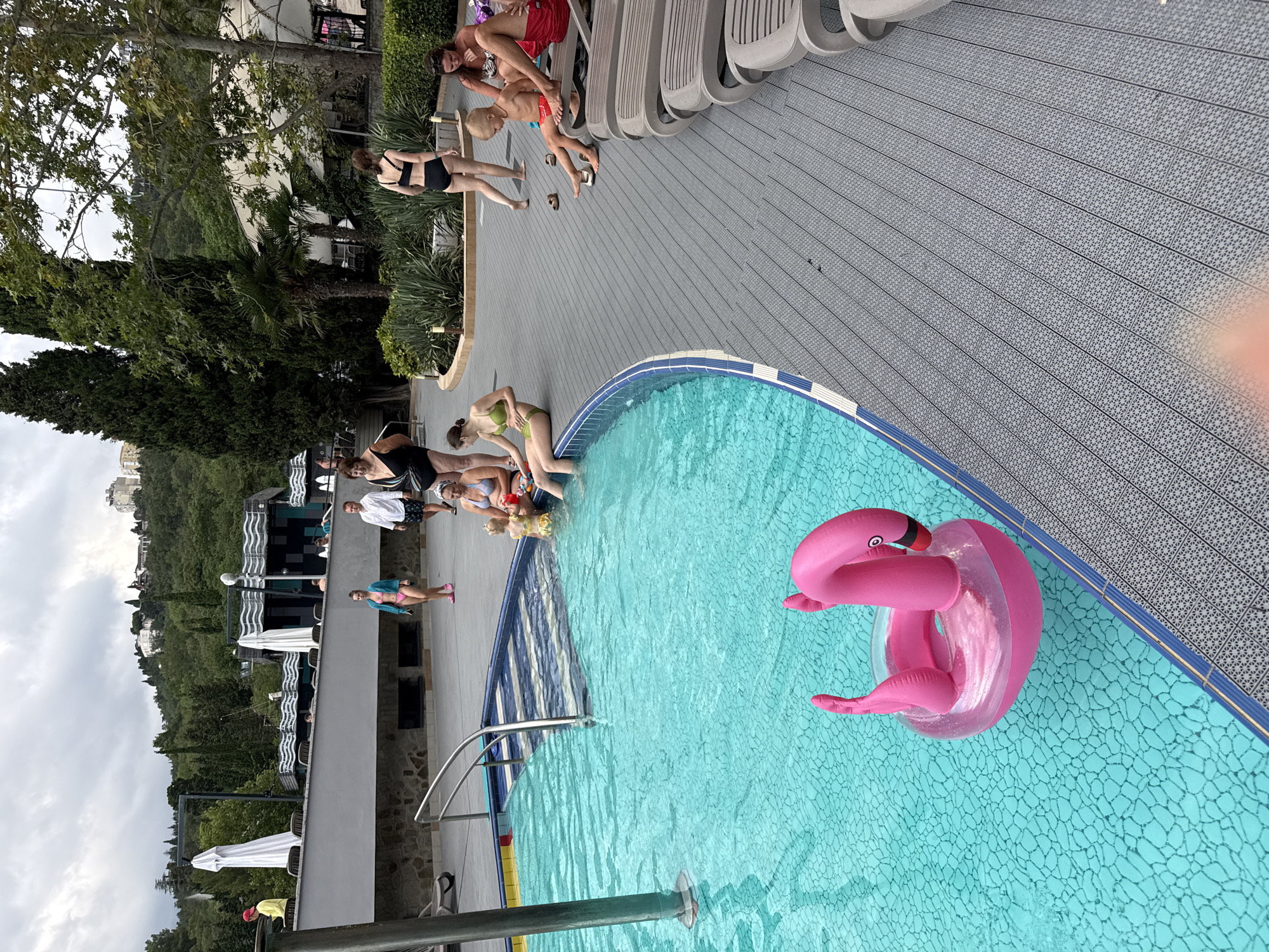 This image depicts a poolside scene at a resort in Massandrovskyi Poselkovyi Radai, Ukraine. In the foreground, a bright pink inflatable flamingo floats in a clear turquoise swimming pool. The pool has a tiled bottom and a blue and white patterned ledge. To the right of the pool, a grey, patterned deck area is lined with several beige lounge chairs, some of which are occupied by people.
In the mid-ground, several individuals are visible. Near the pool's edge, a woman in a green bikini is seated, her legs in the water. Further back, a man and woman in swimsuits are talking, and a young child is sitting between them. To the right, a woman in a black bikini walks away from the pool. A group of people are gathered near the lounge chairs, including a man in red swim trunks, a man in a yellow shirt, and a woman in a black and white swimsuit.
The background shows lush green trees and vegetation covering a hillside. Buildings, some appearing to be part of a hotel or resort complex, are visible on the slope. The sky is overcast and cloudy. The overall atmosphere suggests a leisurely day at a vacation resort.