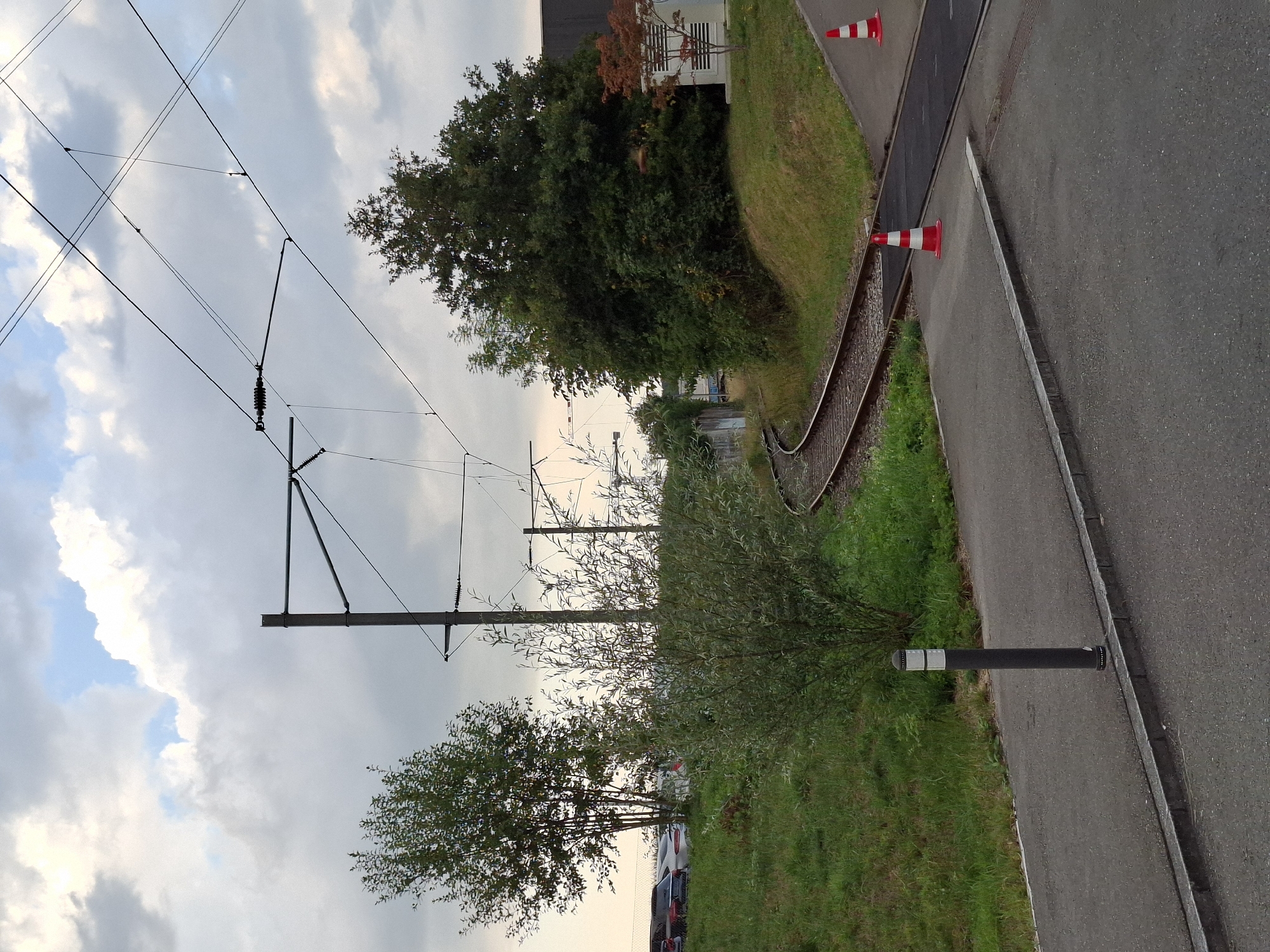 The image depicts an outdoor scene in Rothenburg, Switzerland, featuring a curved railway track that transitions to a crossing. The sky is partially overcast with clouds. In the foreground, a paved path runs alongside a grassy embankment. A dark bollard with reflective bands stands on the path.
To the left of the railway tracks, a grassy slope is visible, with several cars parked in the background. A medium-sized tree with green foliage is situated on this slope.
The railway tracks themselves are made of metal rails and wooden sleepers, with gravel ballast. The tracks curve to the right and lead to a crossing point.
On the right side of the image, beyond the railway tracks, is another paved area with two orange and white striped traffic cones. Behind the tracks and cones, a dense cluster of trees and bushes forms a green backdrop. A light-colored electrical cabinet with a small, red-leafed tree in front of it is also present. In the distance, a construction crane is visible, suggesting ongoing development or maintenance. Overhead, a complex network of electrical wires, likely for the tram or train, is suspended from poles.