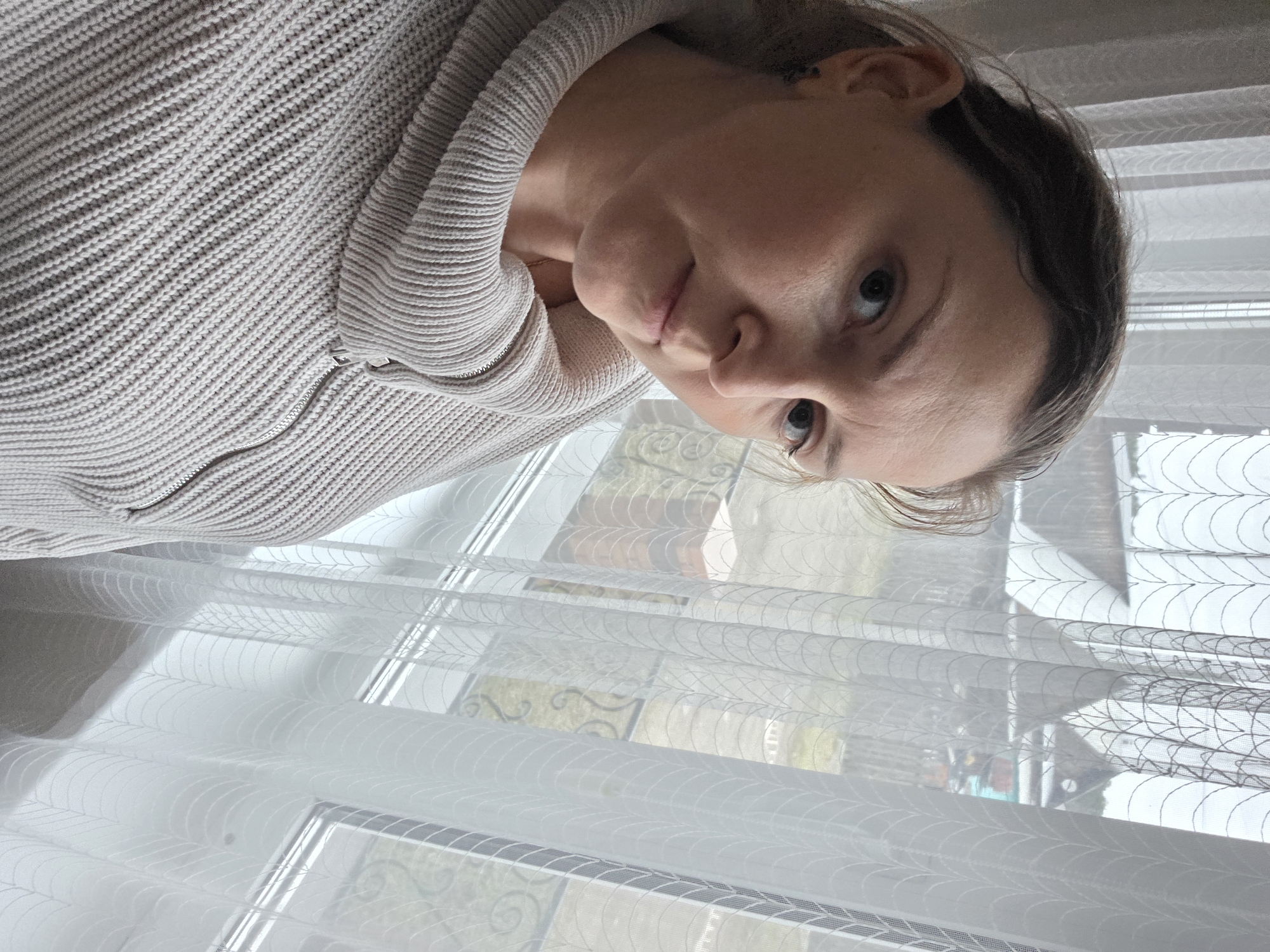 An indoor portrait taken in Chaikovsky, Russia, features a woman looking directly at the camera with a neutral expression. Her head is tilted, and she is wearing a light, possibly oatmeal-colored, knitted sweater with a visible zipper detail on the front. Her dark hair is pulled back, and her eyes are a dark color.
The woman is positioned in front of a window, which is largely covered by sheer white curtains. These curtains have a delicate, repeating pattern of curved lines or leaf-like motifs. Through the translucent fabric and windowpanes, faint architectural shapes, possibly parts of buildings or balcony railings, and diffused outdoor light are visible, suggesting it is daytime. The lighting is soft and natural, illuminating the woman's face and the interior space. No discernible text is visible in the image.