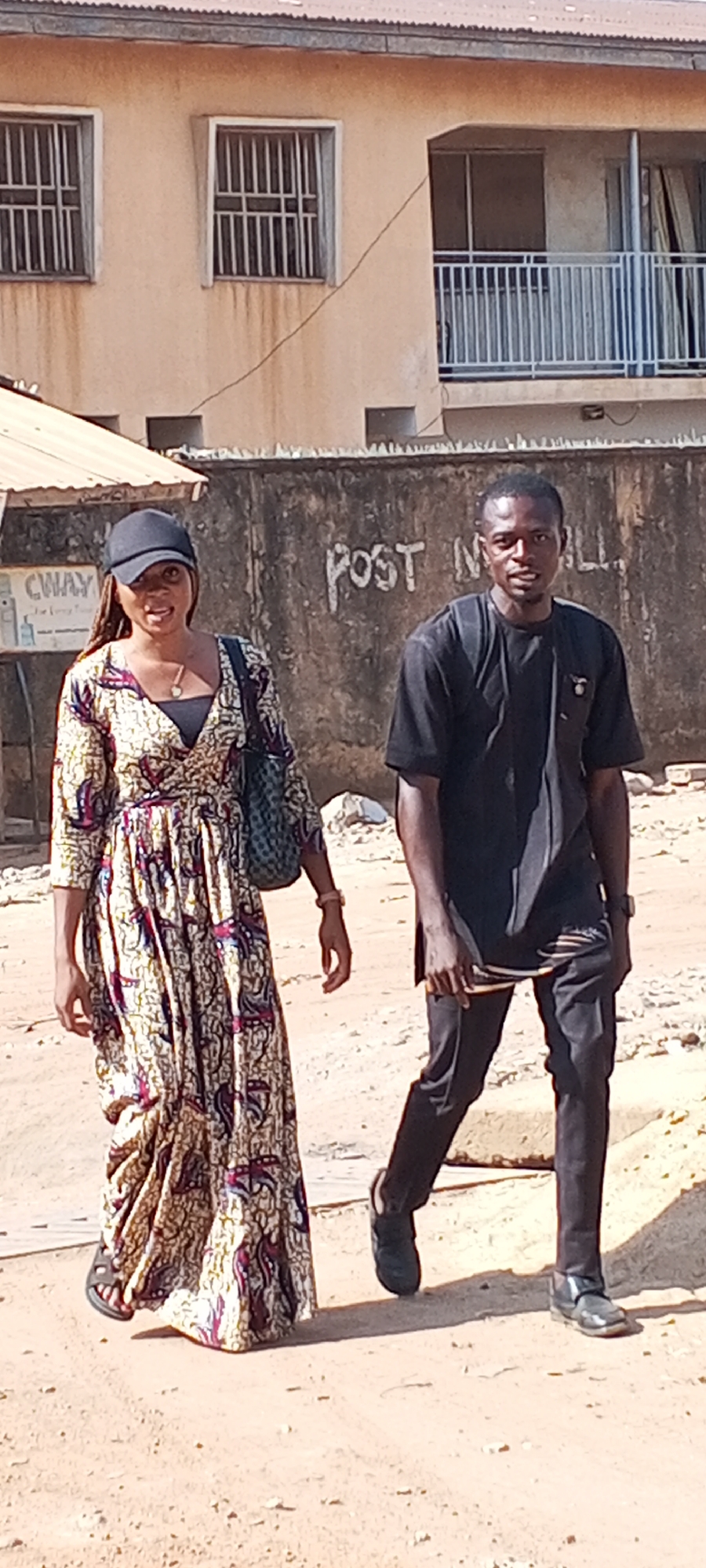 The image captures a daytime scene in what appears to be Sabon Tasha, Nigeria. A man and a woman are walking on a dusty, unpaved street. The woman is on the left, wearing a patterned long dress, a black cap, and carrying a shoulder bag. The man, on the right, is dressed in black attire.
In the background, there's a weathered building and a section of a wall with graffiti partially visible, reading "POST IN ALL". The architectural style of the buildings and the general environment suggest an urban setting, possibly a residential or commercial area in Sabon Tasha.