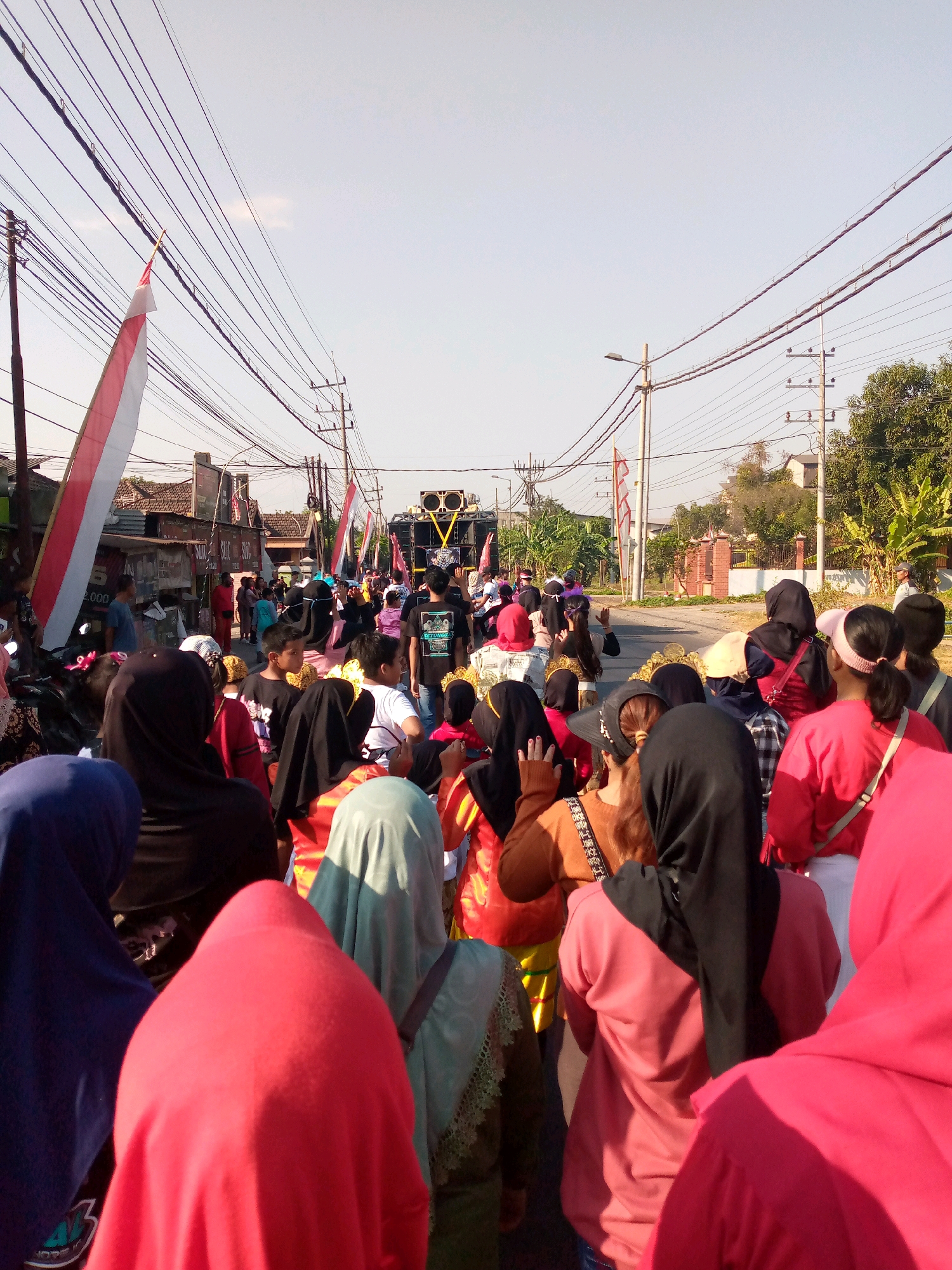 A group of people, many wearing headscarves, are gathered on a street in Sumberwono, Indonesia. The scene appears to be a procession or parade, with a large speaker system on a truck visible in the distance. Indonesian flags are displayed, suggesting a national celebration or event. The street is lined with buildings and trees, and overhead power lines are prominent. The sky is clear and blue, indicating it is daytime. The individuals in the foreground are facing away from the camera, looking towards the procession.