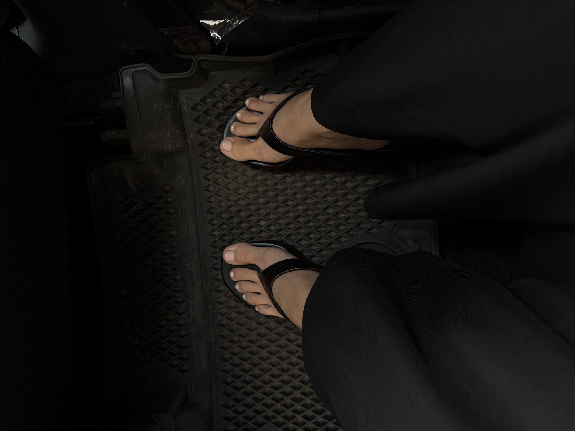 The image shows a close-up view of a person's feet wearing black flip-flops and black pants, resting on a black textured car mat. The scene appears to be inside a vehicle, with the camera angled downwards. The lighting is dim, suggesting an indoor or low-light environment. No specific location within Moscow, Russia, can be identified from the visual information presented in the image.
