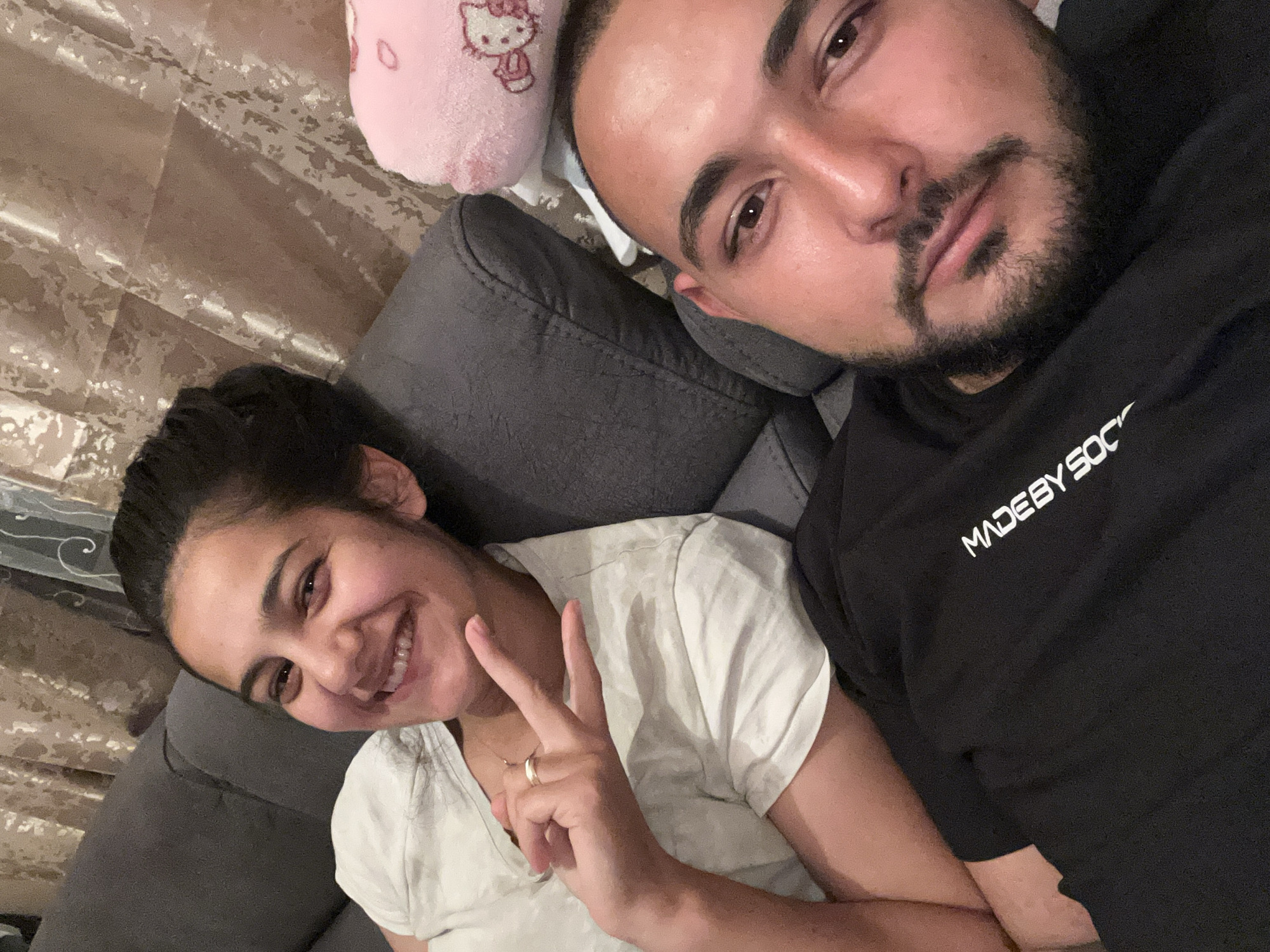 A selfie photograph shows a man and a woman lying on a grey sofa with a pink Hello Kitty plush toy in the background. The woman, positioned on the left, smiles and makes a peace sign with her right hand. She is wearing a white V-neck shirt and has dark hair pulled back. The man, on the right, looks at the camera. He has dark hair, a beard, and is wearing a black hoodie with white text, part of which reads "MADEBY SOC". The background includes a curtain. The specific location in Razhevo Konare, Bulgaria cannot be determined from the provided image.