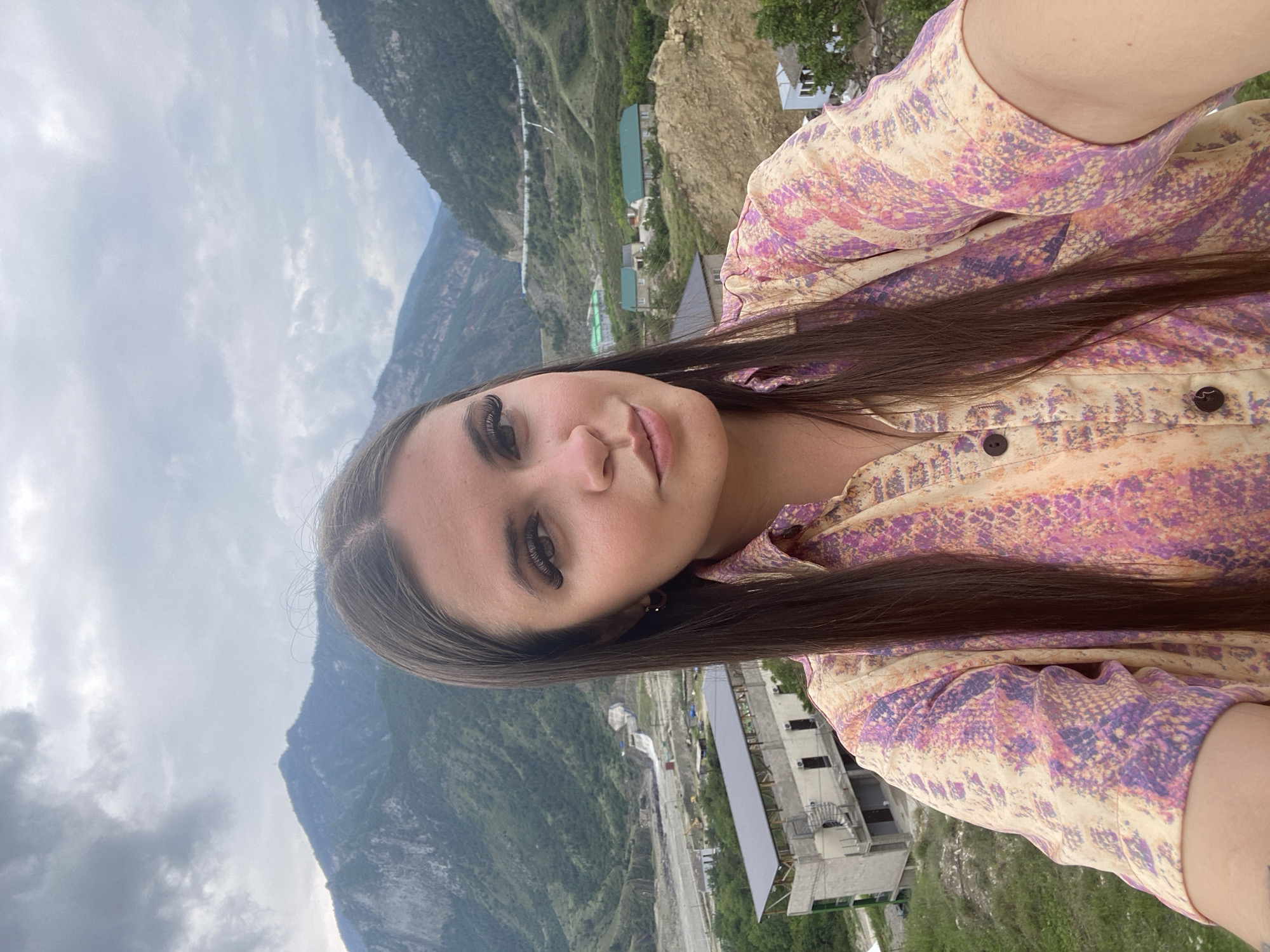 A medium shot shows a young woman with long, dark hair and dramatic makeup taking a selfie in an outdoor environment. She is wearing a colorful, patterned shirt with shades of pink, purple, and yellow. Her expression is slightly pouty. In the background, a mountainous landscape is visible with some buildings and infrastructure, suggesting a semi-rural or developing area. The sky is overcast. The specific location of Chalda, Russia, is not identifiable from the visual elements of the image alone.