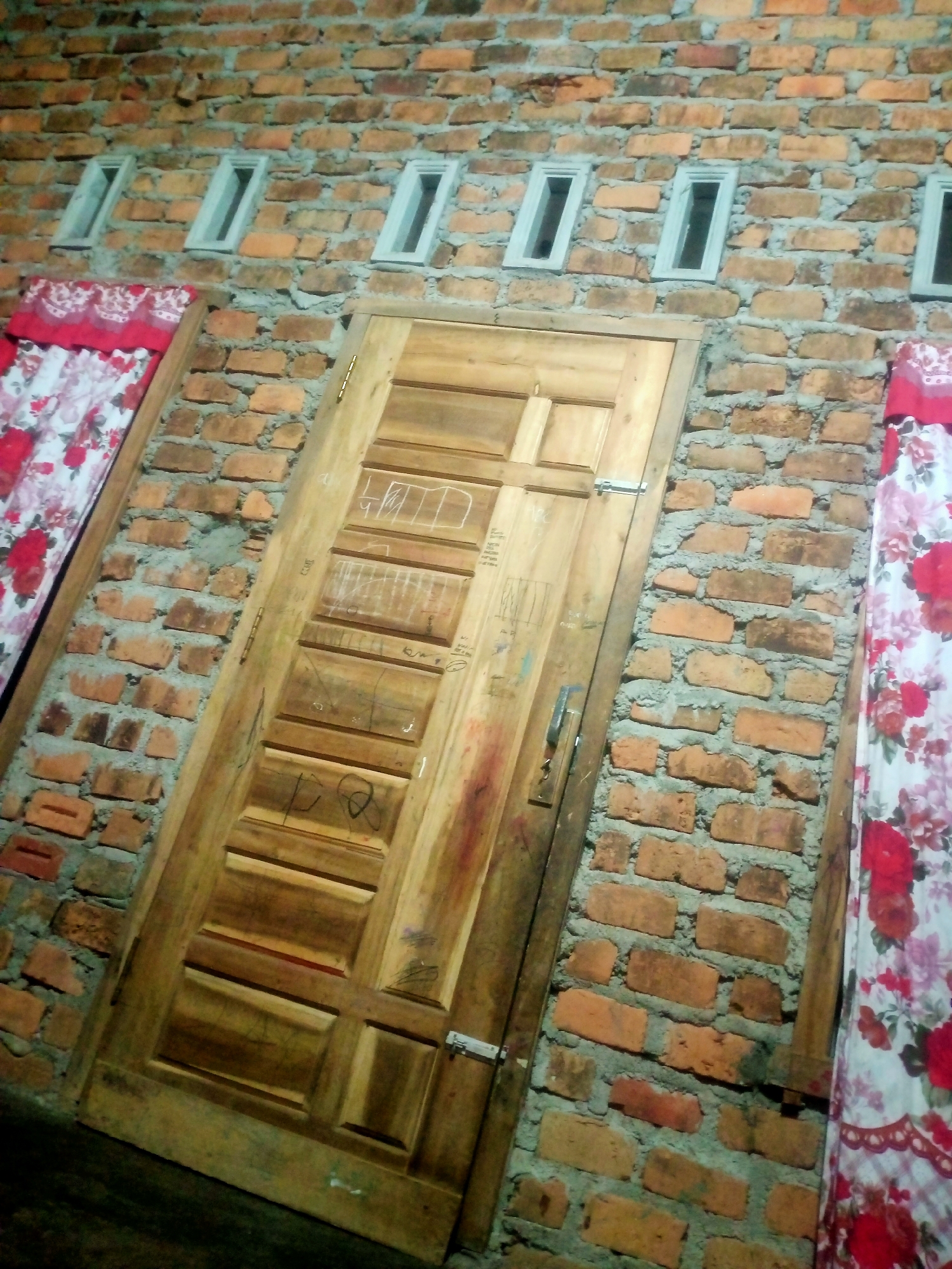The image depicts a wooden door set against a brick wall. There are several small rectangular windows above the door. To the left and right of the door, floral curtains are visible. The brickwork appears unfinished, with visible mortar. The door itself has paneling and a silver handle and latch. Some faint markings or graffiti are present on the door. The overall scene is outdoors, with the lighting suggesting it might be evening or nighttime. The context of Nusa Agung, Indonesia, suggests this is likely a residential exterior.