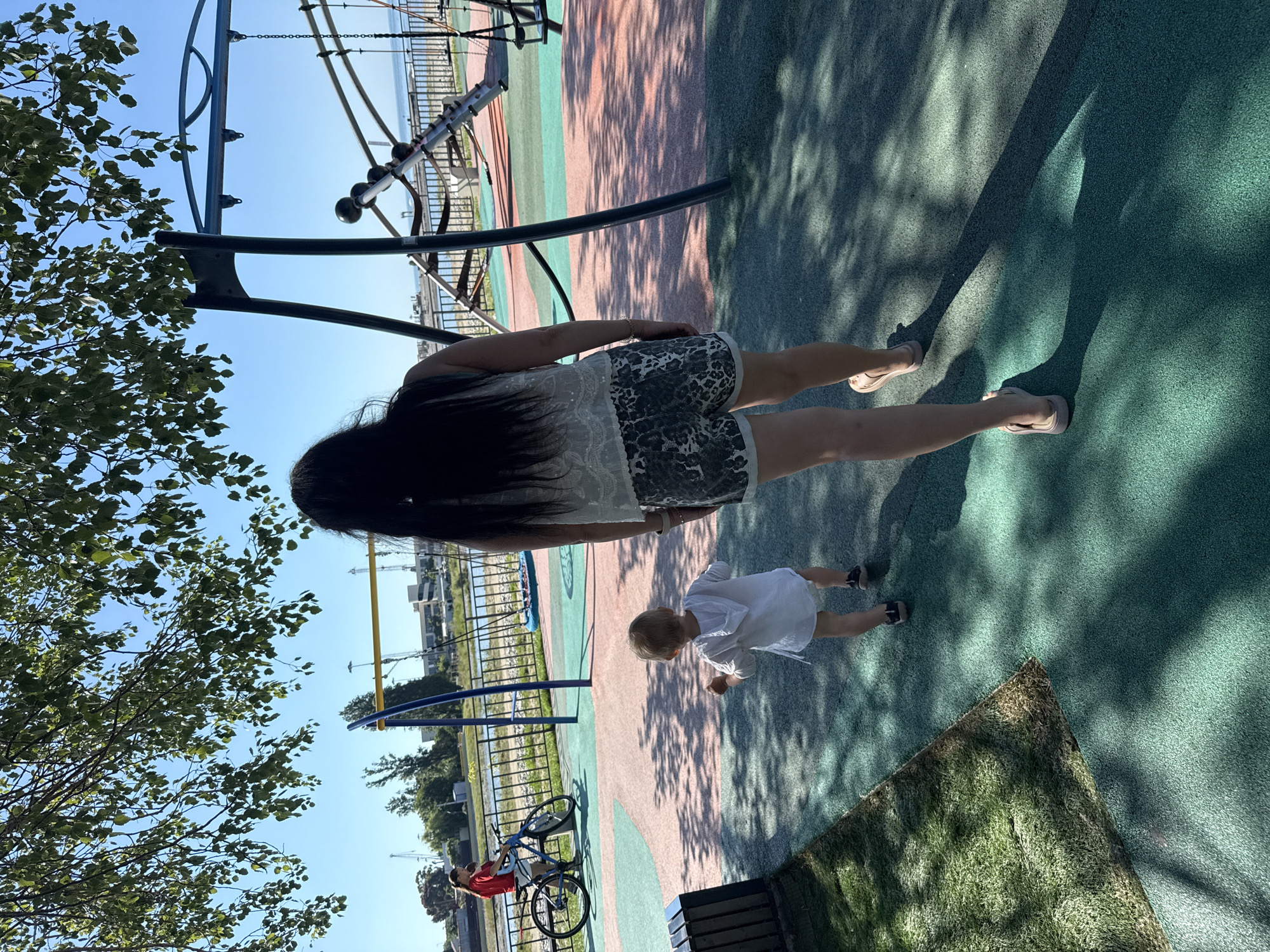 The image depicts a playground scene on a sunny day in Sochi, Russia. In the foreground, a woman with long dark hair walks away from the camera, wearing a light-colored, sleeveless top and patterned shorts. She is followed by a young child, seen from behind, who is wearing a white hooded shirt and dark shoes. The child appears to be walking towards the right of the frame.
The playground features a soft, multi-colored surface, with areas of green, pink, and beige. The background reveals playground equipment, including swings and a climbing structure. Beyond the playground, a body of water, likely the Black Sea, is visible, with boats and industrial structures on the horizon. The scene is partially shaded by the leaves and branches of a large tree in the upper left corner of the image. In the middle ground, to the left, a woman on a bicycle is also visible, with a child sitting on the handlebars. The overall impression is one of a recreational area within a coastal city.
