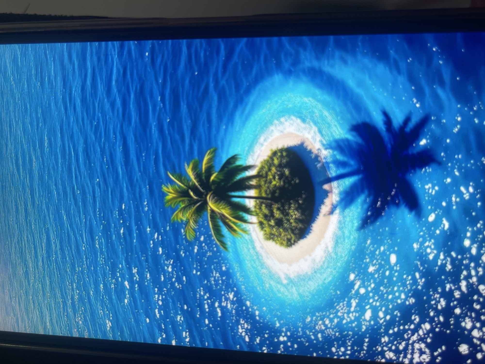 The image shows an aerial view of a small, uninhabited tropical island in the middle of a vibrant blue ocean. The island is round with a white sandy beach and covered in lush green vegetation, dominated by a single tall palm tree. The water surrounding the island is clear and shallow near the shore, creating a bright turquoise halo, which deepens to a rich blue further out. Whitecaps and foam are visible on the surface of the water, suggesting gentle waves. A dark shadow of the palm tree is cast on the sandy beach and extends into the water. The overall impression is one of serene isolation and natural beauty. The image does not contain any elements that suggest a location in Bishkek City, Kyrgyzstan, as it depicts a tropical environment.