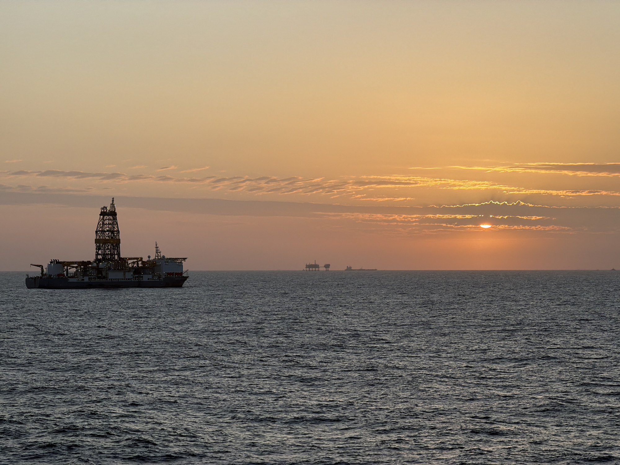 The image depicts a seascape at sunset. In the foreground, a large drilling ship is visible on the left side of the frame. In the distance, there are silhouettes of what appear to be oil platforms or other maritime structures on the horizon. The sun is partially obscured by clouds, creating a warm, golden glow across the sky and water. The environment consists primarily of the ocean and the sky, with no land visible. The location is the city of Unknown, Unknown.