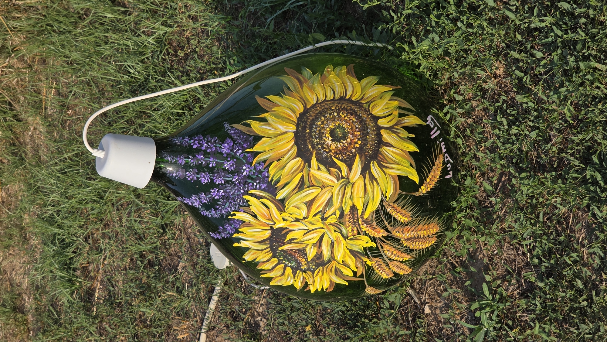 A green glass demijohn, painted with sunflowers, lavender, and wheat stalks, sits on grass. A white electrical cord emerges from a grey cap on the demijohn's opening. The demijohn is decorated with the name "Ludmilla" in pink cursive lettering. The scene is outdoors, with green grass covering the ground, suggesting a natural or garden setting. The image does not contain any specific visual cues to pinpoint a location within the city of Tuscania, Italy.
