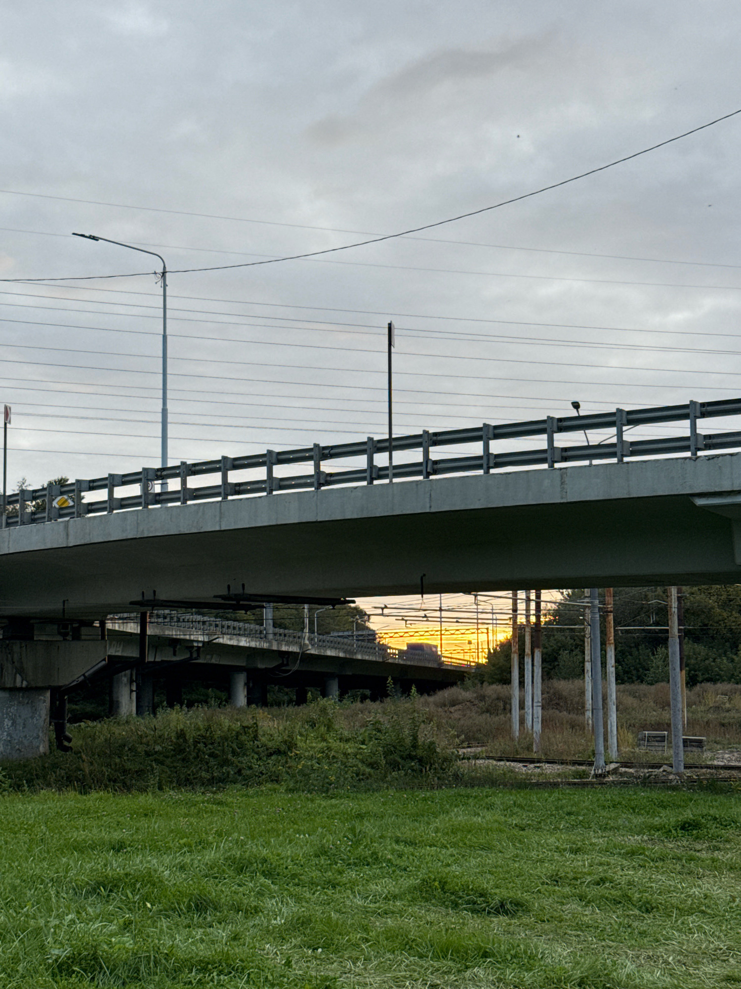 The image shows a two-level overpass crossing a grassy field under a cloudy sky. The upper level of the overpass is a concrete bridge with a metal guardrail and lights. Below it, another overpass is visible with concrete pillars supporting it. The lower overpass is partially obscured by vegetation and appears to be part of a railway or highway system. In the background, trees and utility poles are present. The sky is overcast with a hint of sunset visible through a gap in the clouds, casting a warm glow. The foreground is a field of green grass. This scene is likely located in Yaroslavl, Russia, as suggested by the presence of overhead power lines and the style of the overpasses, which are common in Russian urban infrastructure.