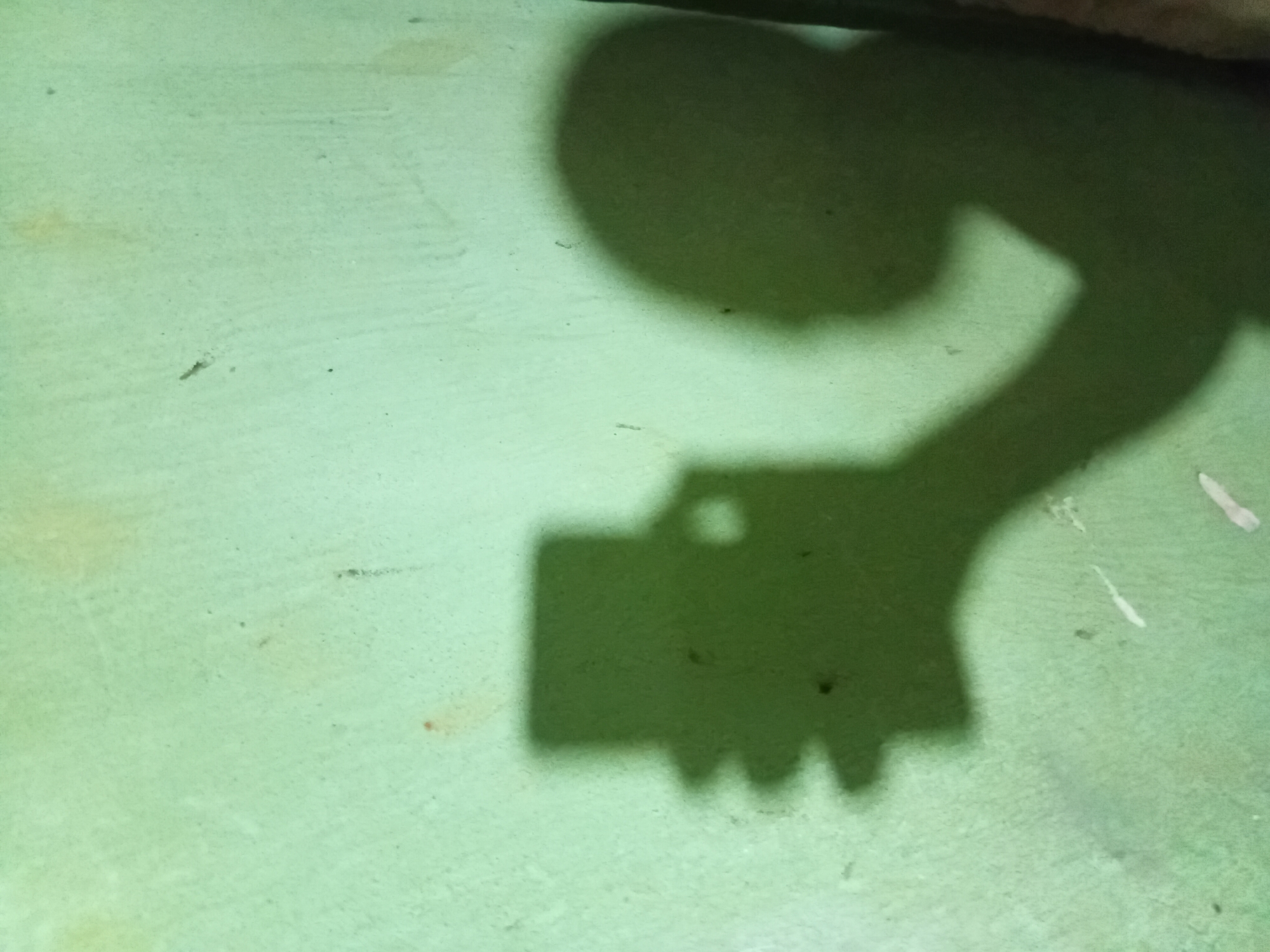 This image captures the shadow of a person holding a smartphone, casting a silhouette against a light green, textured wall. The shadow on the left depicts a hand holding a rectangular object, likely the phone, with fingers splayed. To the right, the shadow of a head and upper body is visible, suggesting the person is taking a selfie. The wall itself exhibits minor imperfections, with some lighter patches and small dark marks. The lighting suggests an indoor environment with a light source positioned to create these shadows. The scene is likely in a residential or public building in Nkpor, Nigeria, given the context.