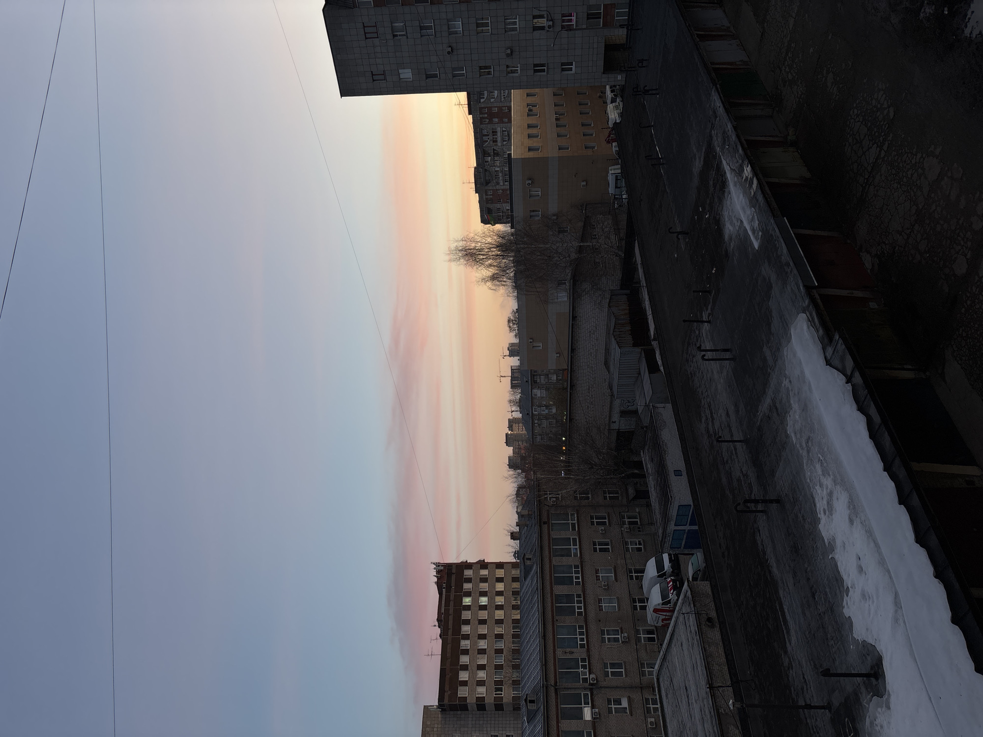 The image depicts a cityscape in Perm, Russia, captured from an elevated vantage point, likely a rooftop. The foreground shows a flat, dark rooftop covered in places with remnants of snow or ice. Metal protrusions, possibly supports or vents, are sparsely distributed across the roof. Adjacent to the roof, the scene unfolds with multiple apartment buildings of varying heights and architectural styles. The buildings are primarily concrete and feature repetitive window patterns, characteristic of Soviet-era construction. Some buildings have air conditioning units attached to the exterior walls. A few bare trees are interspersed among the buildings. In the background, the sky transitions from a light blue at the top to a blend of pink, orange, and yellow towards the horizon, indicating either a sunrise or sunset. Electrical wires stretch across the sky in the upper-left corner of the image. No people are visible.