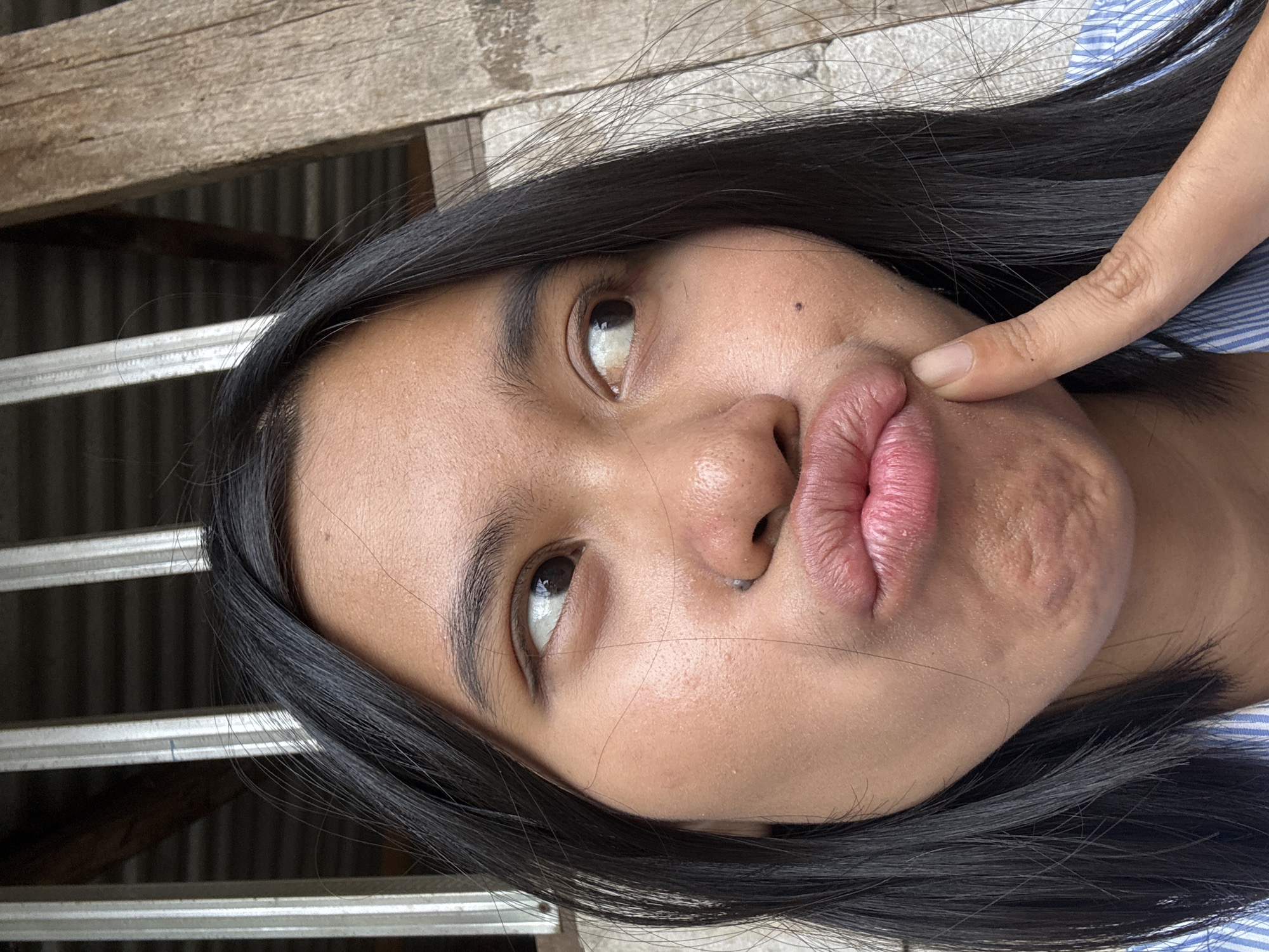 A close-up, low-angle, waist-up shot features a young woman with dark hair and brown eyes. She is looking to her right with her lips pursed, and her right index finger is touching her left cheek. She is wearing a blue and white striped shirt. The background consists of corrugated metal siding and wooden planks. The scene appears to be outdoors in East Jakarta, Indonesia, with the rustic architectural elements suggesting a residential or semi-rural setting.