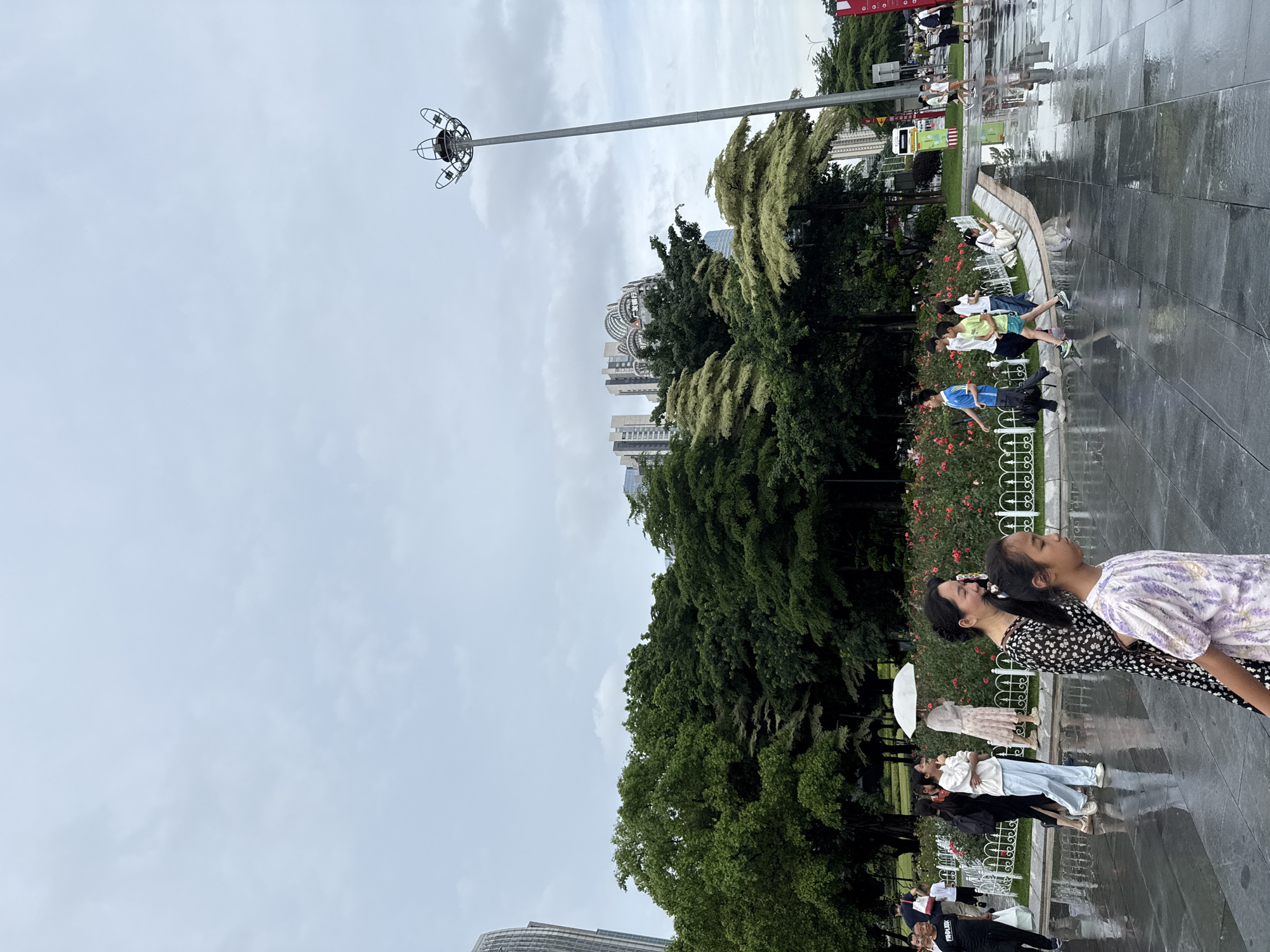A medium shot captures a street scene in Tianhe District, China, under an overcast sky. The wet, reflective pavement suggests recent rainfall. In the foreground, a woman in a floral dress walks with a young girl in a purple and white patterned top. To their left, a line of people walks along a white picket fence bordering a flower bed with red roses. Among them, a woman holding a white umbrella is visible. Behind the fence, lush green trees form a dense canopy. In the background, modern buildings rise above the trees, with a tall, distinctive street lamp on the right. Several individuals are seen walking and interacting on the wet pavement, some carrying bags.