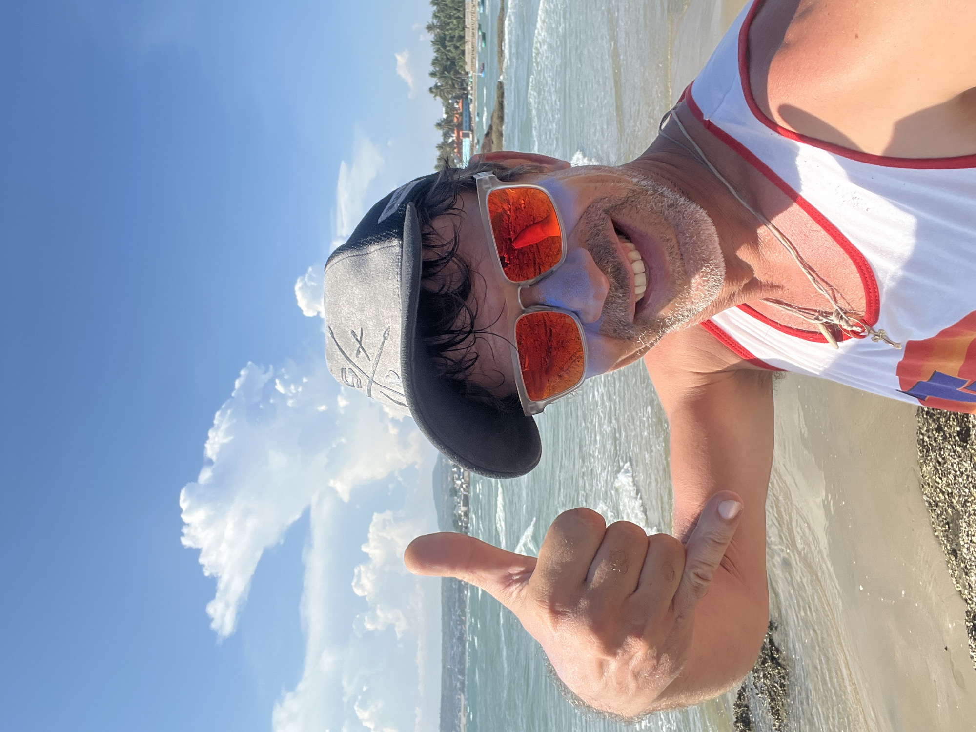 A man is taking a selfie on a beach in Mũi Né, Vietnam. He is wearing a grey baseball cap, red-tinted sunglasses, and a white tank top with red trim. He has a beard and appears to have sunscreen on his cheeks. The man is smiling and giving a "shaka" hand gesture. The background shows the ocean, sand, and some palm trees. The sky is blue with some white clouds. There are a few boats visible in the water.