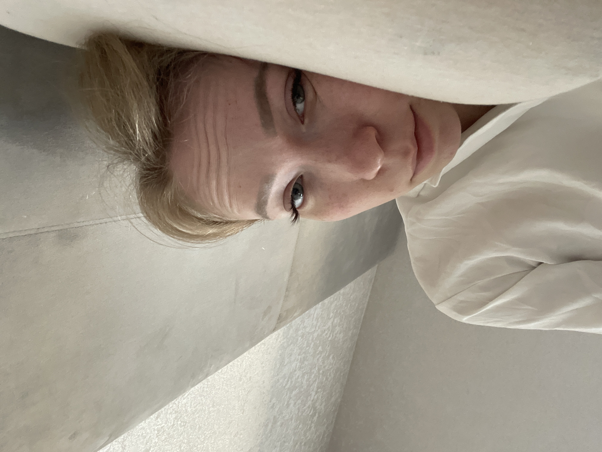 A close-up, upside-down perspective shows a fair-skinned individual with blonde hair, appearing to be indoors. The person's face is partially obscured by what seems to be a pillow or blanket, and they are wearing a white garment. The background consists of what looks like a wall or another piece of furniture with a neutral, light color. No features indicate location or time of day, nor are there any identifiable landmarks or objects to ascertain that the photo was taken in Perm, Russia.