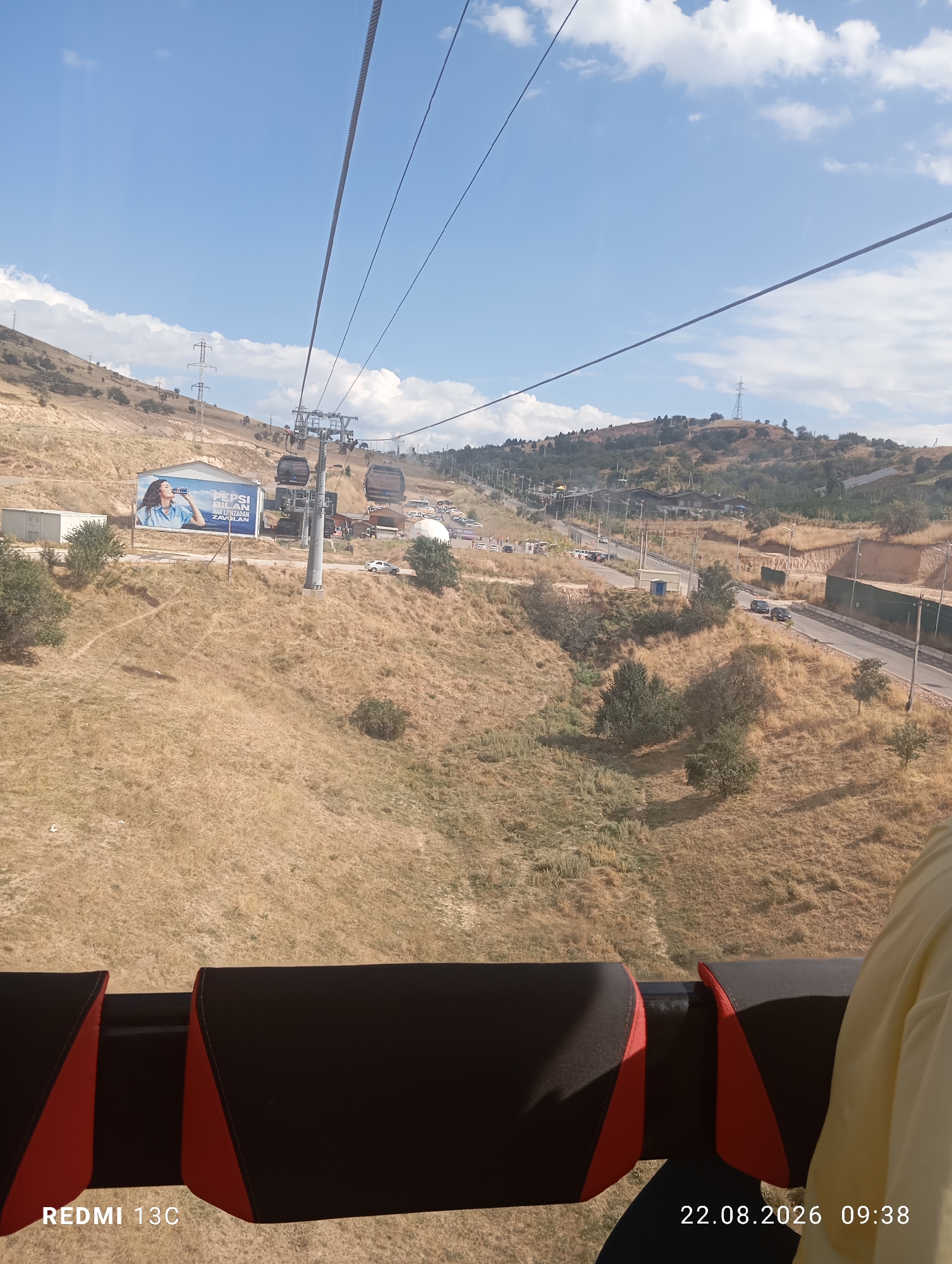 The image shows a view from a cable car ascending a dry, grassy hillside under a partly cloudy blue sky. In the foreground, the red and black seats of the cable car are visible. Further down the hillside, a Pepsi advertisement is displayed on a billboard. Several cable cars are suspended from overhead cables, moving along the slope. A road with cars and a small building are visible to the right. The general landscape is arid, with sparse vegetation and rolling hills. The location is identified as Amirsay, Uzbekistan. The date and time stamp at the bottom right corner reads "22.08.2026 09:38".