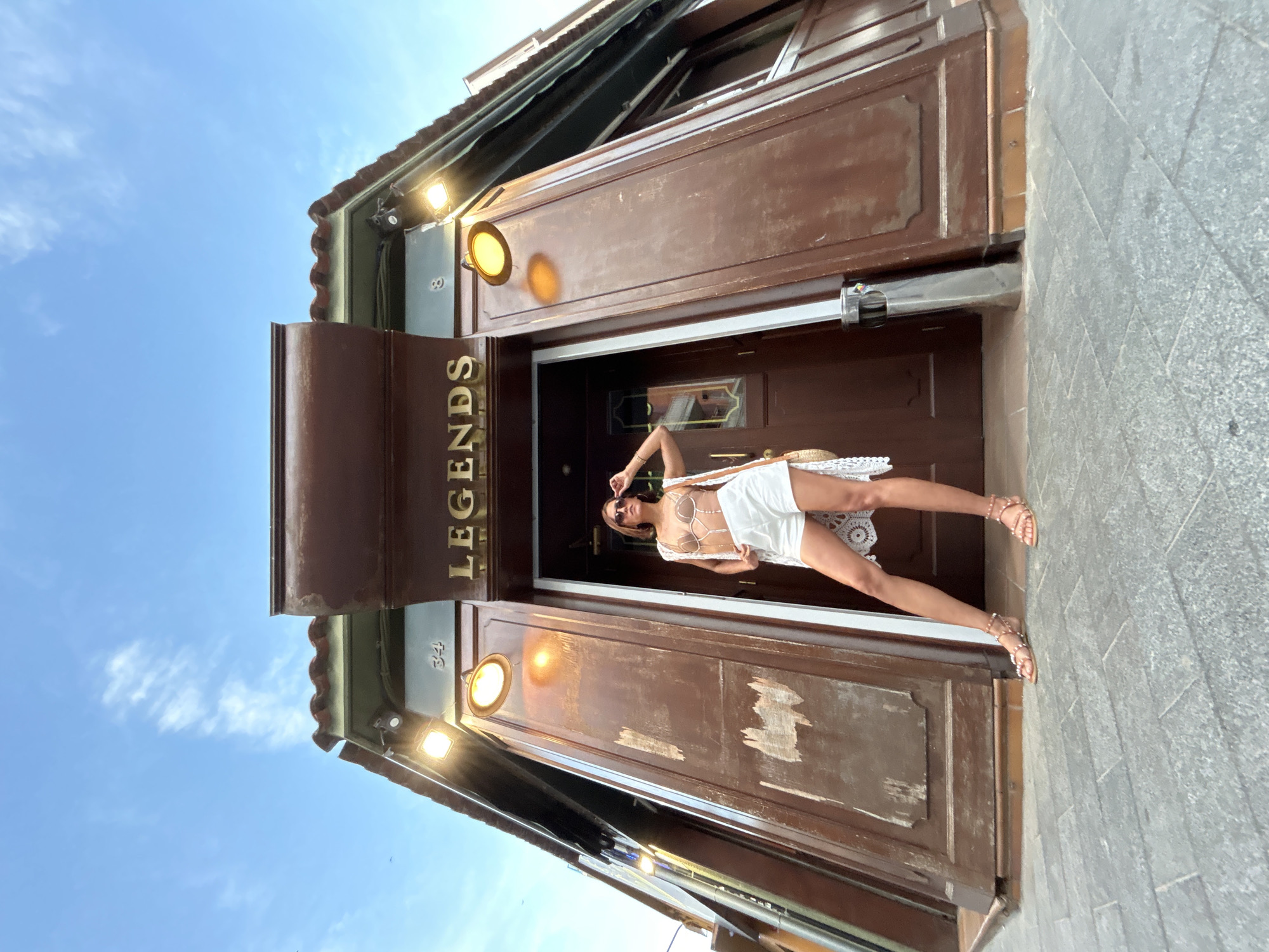 This low-angle photograph captures a female subject standing in front of a building entrance identified by the name "LEGENDS" in Calella, Spain. The subject, a young adult female, stands centrally, facing the camera with a slight upward gaze. She has dark hair, wears sunglasses pushed onto her head, a light brown, possibly beaded or sequined, halter-neck bra-top, white high-waisted shorts, and a white open-knit or crocheted long vest. Her right hand is on her hip, and her left hand is raised towards her sunglasses. She wears light-colored, multi-strapped sandals.
The building entrance is constructed of dark brown wooden panels, exhibiting wear with visible scuffing and peeling paint. Above the double door, the word "LEGENDS" is displayed in gold, capitalized letters. Flanking the entrance are two light fixtures, one on each side, emitting a warm yellow glow. Above these lights, the numbers "34" and "8" are visible on the left and right, respectively, inscribed on a lighter-colored band beneath a tiled roof overhang. A modern, cylindrical, silver trash receptacle is partially visible to the right of the subject's left leg, positioned against the building. The ground is paved with rectangular grey cobblestones or tiles. The sky above is clear blue with sparse, wispy white clouds.