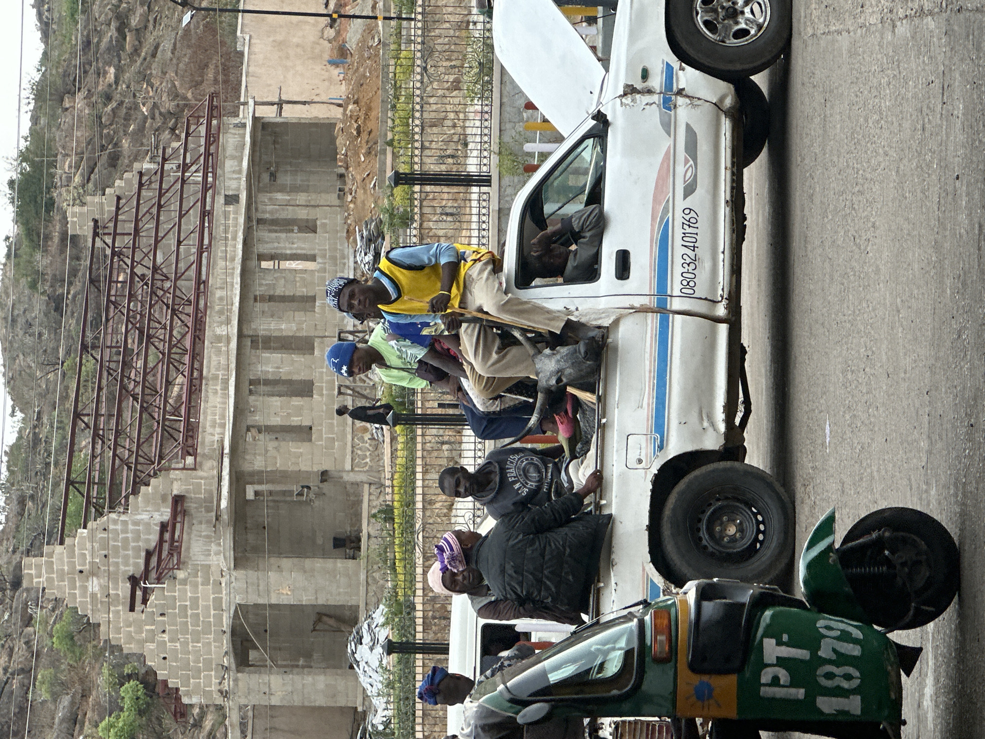 This medium shot captures a vibrant street scene, likely in Jos, Nigeria, showing a crowded white utility vehicle and a partially visible green tricycle against a backdrop of a rocky hillside and construction.
The central subject is a white, weathered vehicle, possibly a pickup truck or minibus, with its hood open. It is heavily laden with people, many of whom are seated or standing on its exterior. On the passenger side, a man in a yellow vest and patterned head covering sits, holding a stick. Below him, another man wears a blue cap. Several other men are visible on the vehicle, including one in a black hoodie with "SAN FRANCISCO" and "CALIFORNIA" emblazoned on it, and another in a black jacket and a pink/purple head covering. The vehicle shows signs of age, with peeling paint and rust, and features blue and brown stripes along its side. A spare tire is visible at the rear.
In the foreground, the front and side of a green tricycle, commonly known as a Keke Napep, are visible, partially obscured by the white vehicle. It has yellow accents.
The background reveals a rugged, rocky hillside with sparse vegetation. Below the hillside, concrete block structures in various stages of construction are visible, some with exposed rebar. A metal fence or gate runs in the mid-ground. Overhead, utility lines traverse the sky.
The scene depicts a bustling, everyday activity, likely public transportation. The vehicles appear well-used, and the people are casually dressed. The lighting suggests daytime, possibly under an overcast sky.
Visible text includes a phone number "080-32 401769" on the side of the white vehicle and "PT- 1879" on the green tricycle, which could be a registration number.
