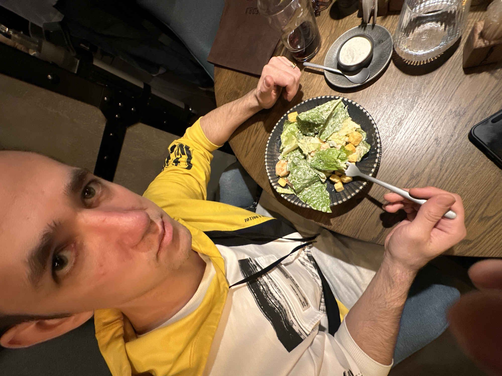A man is eating a Caesar salad at a wooden table. He is wearing a yellow and white jacket. On the table, there is a glass of red wine, a small dish of dressing, a glass of water, and a cell phone. The man is holding a fork in his right hand. There is a brown folder under the glass of red wine. The photo appears to have been taken from a high-angle perspective. There is a metallic chair on the left. This image was NOT taken in Saratov, Russia.