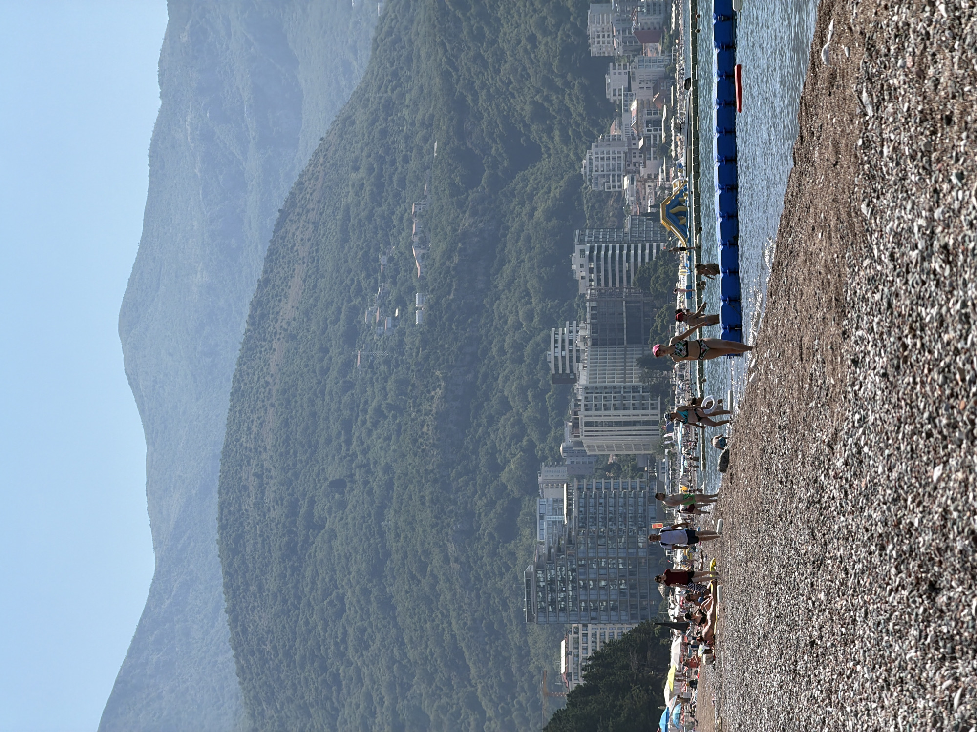 The image depicts a sunny day at a pebble beach in Boreti, Montenegro. In the foreground, the beach is covered in small, multi-colored pebbles and some seaweed. A few people are walking along the shoreline, and some are wading in the shallow water. In the middle ground, the sea is calm, and a blue floating barrier separates the swimming area from the open water. Beyond the beach, a line of modern, multi-story buildings, including hotels and apartments, stretches along the coastline. Behind the buildings, a lush, green mountainside rises steeply, covered in dense vegetation. The sky is clear and blue, with no clouds visible. The overall scene is one of a typical Mediterranean coastal resort on a bright summer day.