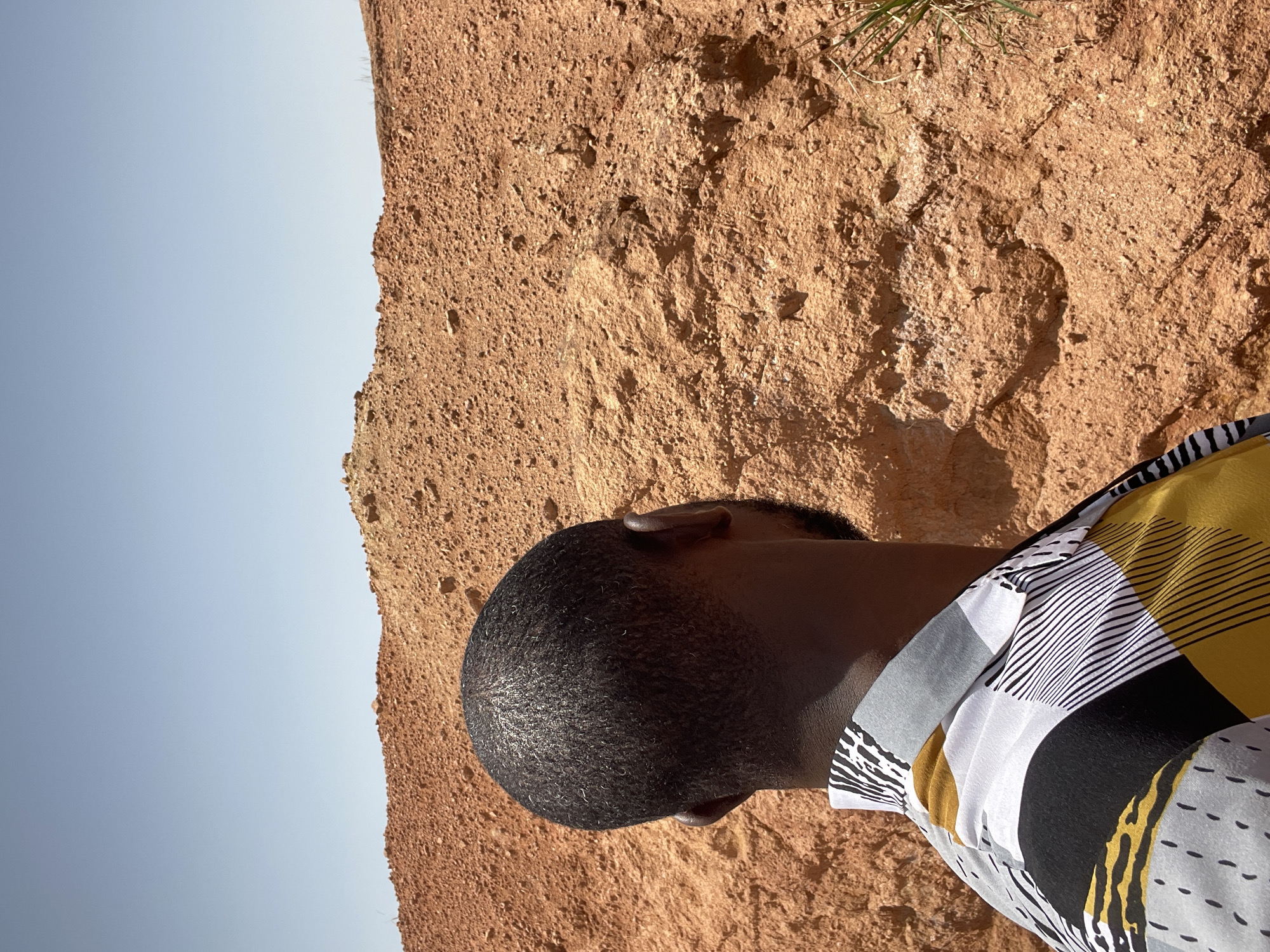 An over-the-shoulder shot captures the back of a Black man's head and shoulders against a backdrop of a dusty, orange-red, rocky landscape. The man is wearing a collared shirt with a bold, abstract pattern of black, white, yellow, and gray geometric shapes and lines. The sky above is a clear, pale blue. The terrain is characterized by uneven, eroded earth and scattered rocks, with a hint of dry grass on the right side of the frame. This scene appears to be an outdoor environment in the city of Jos, Nigeria, given the dry, arid appearance of the land and the clear sky often seen in such locations. The focus is on the man's profile as he looks out at the landscape.