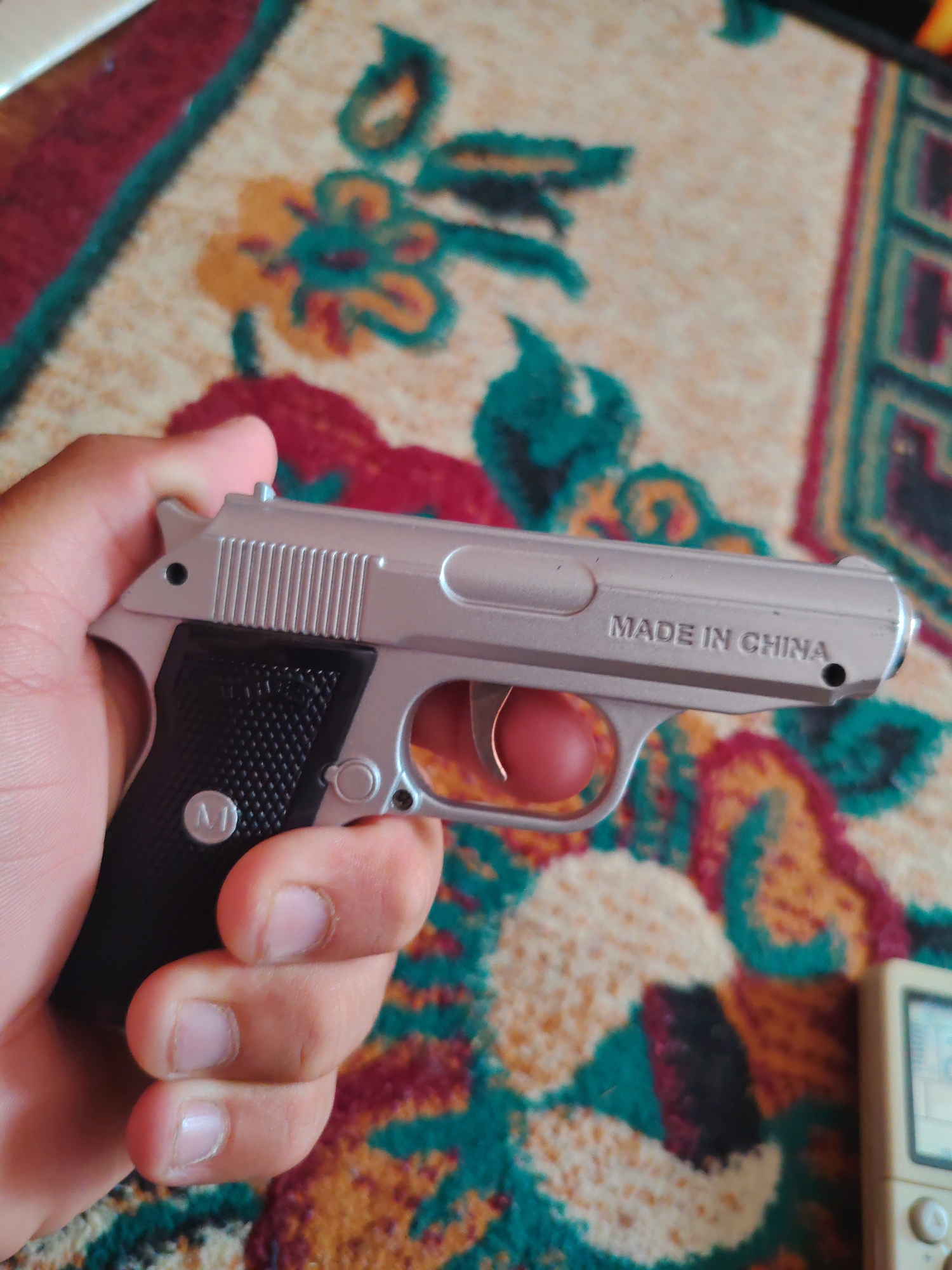 A close-up, high-angle, slightly blurred shot shows a person's right hand holding a silver toy pistol. The pistol has a black grip with a textured pattern and a circular emblem with the letter "M" inside. The slide of the pistol is engraved with "MADE IN CHINA". The background is a colorful, floral-patterned rug. The scene appears to be indoors, possibly in a home, in the city of Khujand, Tajikistan.