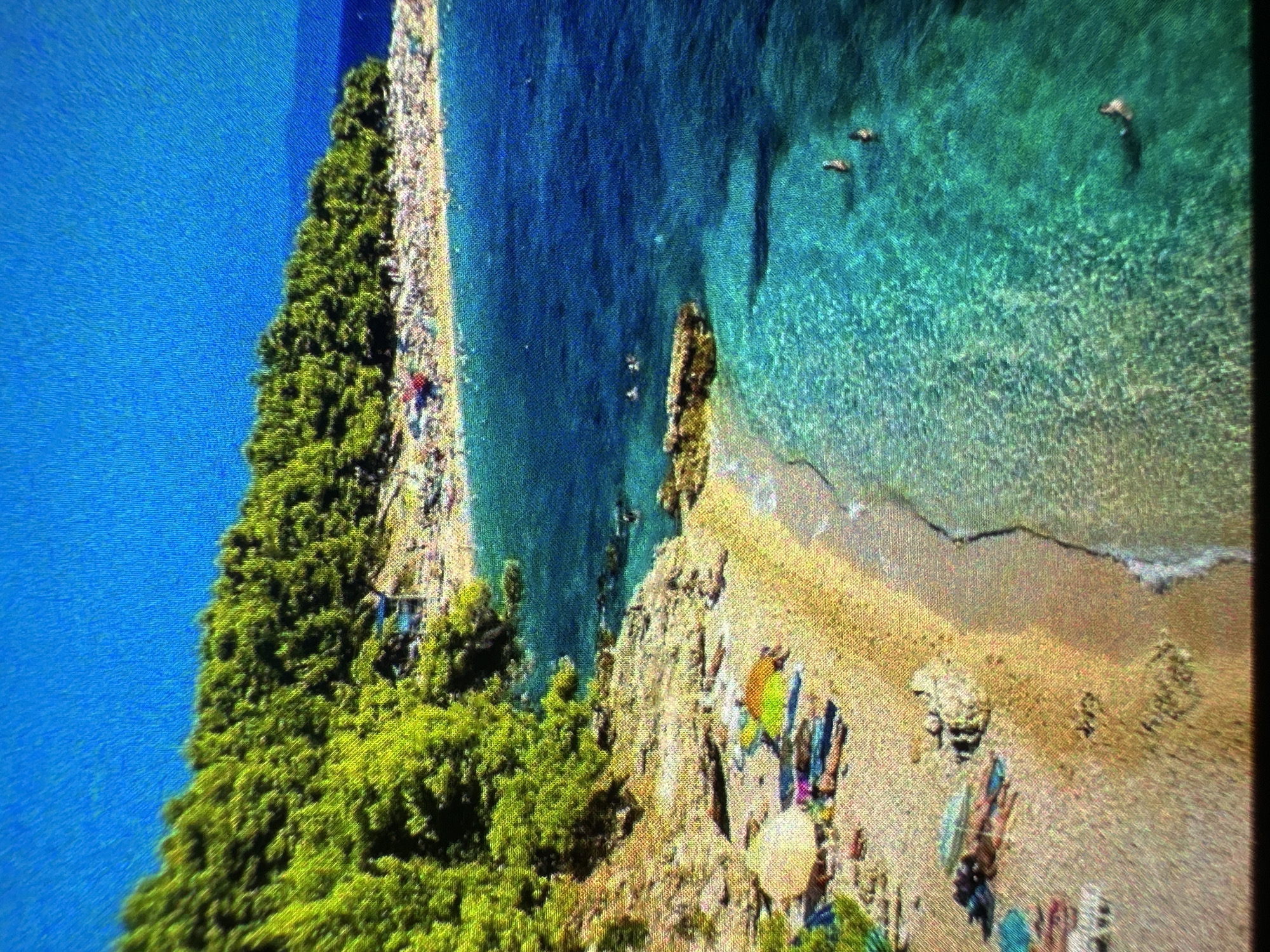 An aerial view captures a sun-drenched beach with turquoise waters. Dense green trees line the upper edge of the frame, with a small cluster of buildings visible nestled among them. Below the trees, a densely populated beach stretches along the coastline, with numerous people sunbathing and wading in the water. Closer to the foreground, a sandy beach area is dotted with beach chairs, umbrellas, and a few individuals. The clear water reveals the sandy seabed below, and several swimmers are visible in the ocean.