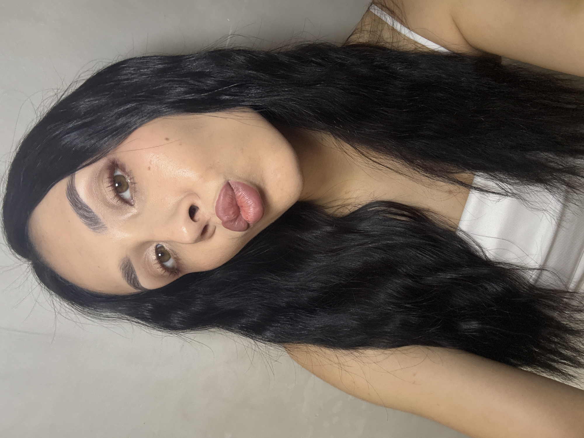 The image is a close-up selfie of a young woman with long, dark, wavy hair and fair skin. She is wearing a white top. Her eyes are open and looking towards the camera, and her lips are pursed in a kissing gesture. She has on makeup, including eyeshadow and lipstick. The background is a plain, light-colored wall. The image was taken indoors. The question about the location in Votkinsk, Russia is not answerable from the image content.