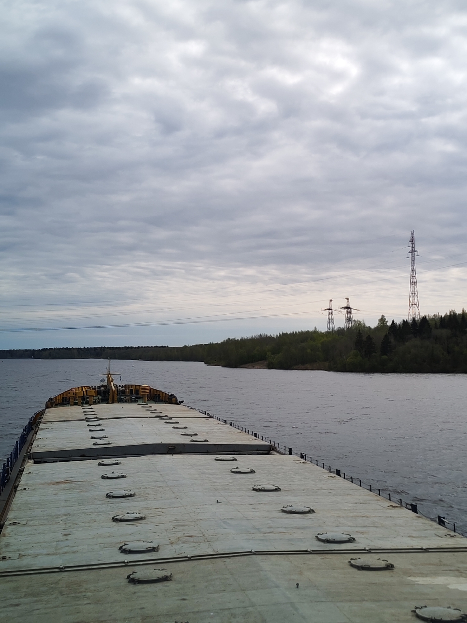 The image presents a view from the deck of a barge on the water, possibly a river or large lake. The deck of the barge is gray and featureless except for multiple circular hatches. The water is gray and slightly choppy. In the distance, a forested shoreline is visible. Three power transmission towers are visible along the shoreline. The sky is overcast with gray and white clouds.
While it is not possible to pinpoint the exact location as Борисово, Russia using just this image, the presence of a wide body of water with forested shores and power transmission towers aligns with the characteristics of rivers and lakes in Russia.