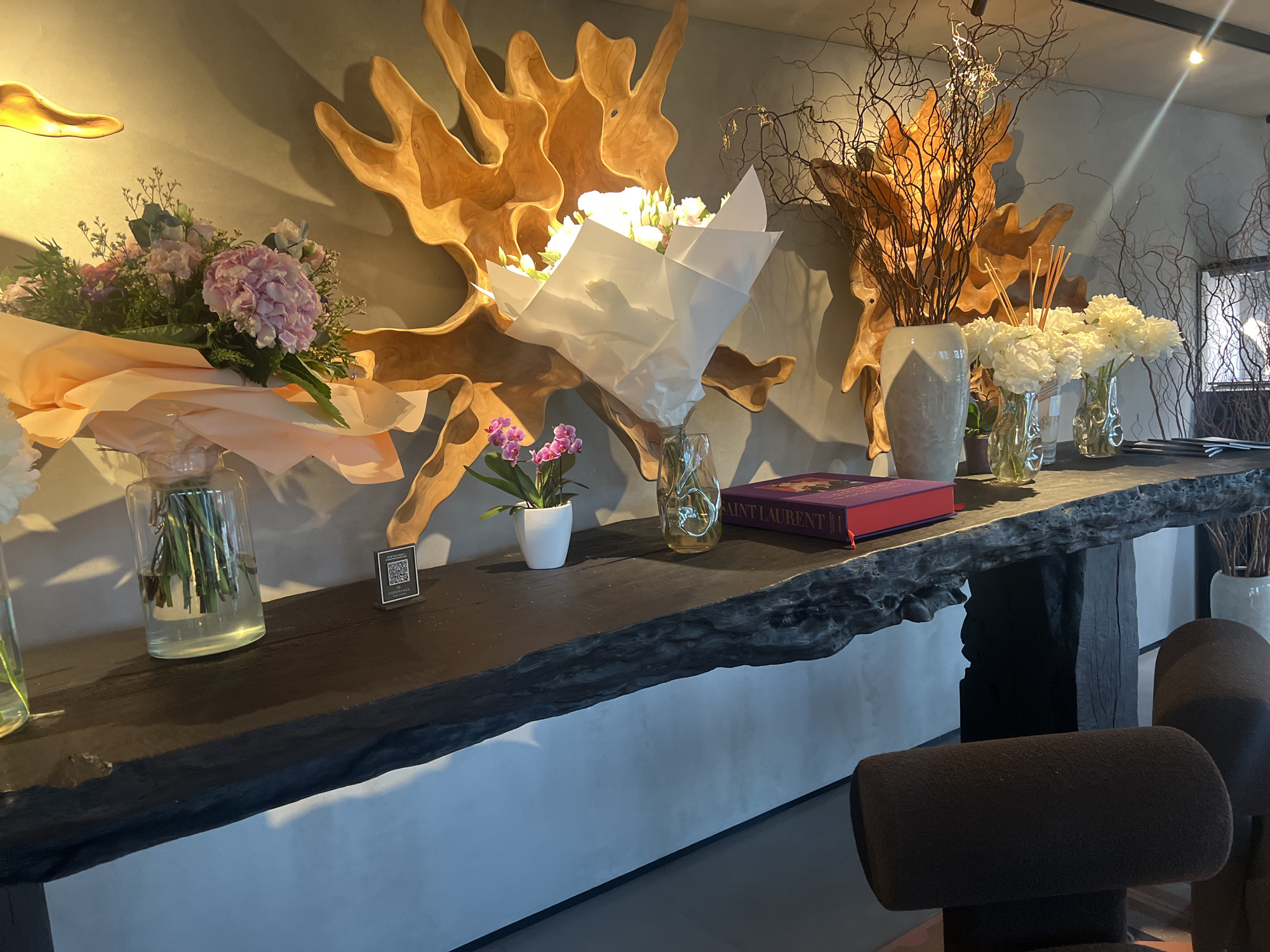 The image depicts a close-up view of a rustic wooden console table in a well-lit interior space. The table is adorned with several floral arrangements and decorative items. On the left, a bouquet of pink and white flowers is partially wrapped in pale orange paper. To its right, a small potted orchid with pink flowers sits in a white pot. Further to the right, a bouquet of white flowers is presented in white paper wrapping. The console table itself is made of dark, rough-hewn wood with an irregular edge, and it rests on a dark base. Behind the table, large, abstract wooden sculptures are mounted on a textured wall. On the table, to the right of the white floral arrangement, are two clear glass vases holding white flowers and some reeds. A thick, dark red book with gold lettering, possibly a Yves Saint Laurent publication, is placed on the table to the right of the vases. The background shows a light-colored wall with shadows cast by the decorative elements. In the foreground, the backs of two dark brown upholstered chairs are visible, suggesting a seating area. The overall impression is one of elegant, natural decor. This scene is located in Užilgiai, Lithuania.