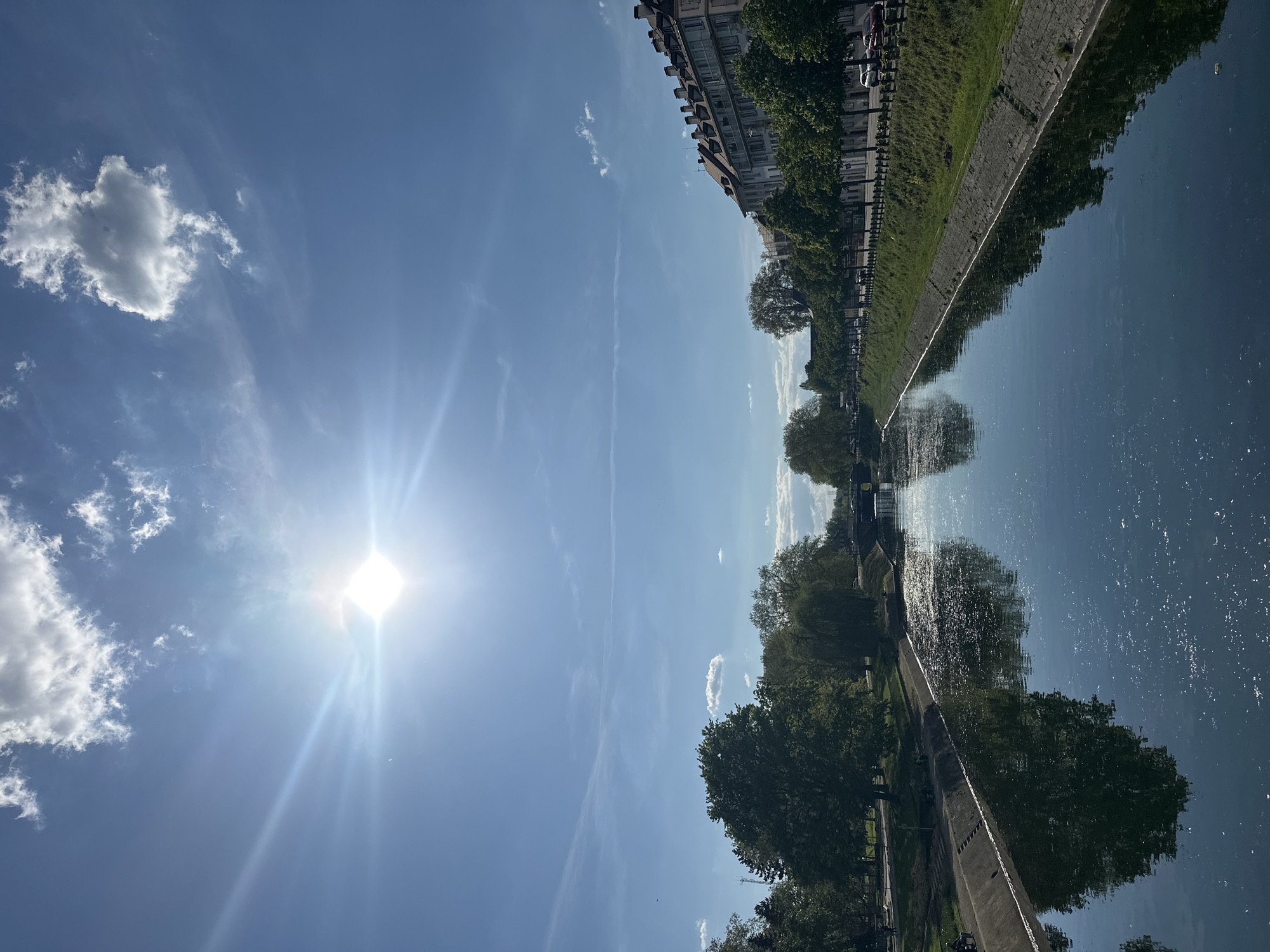 The image captures a canal in Strasbourg, France, on a sunny day. A body of water reflecting the trees, buildings, and sky, runs horizontally across the frame. The sky, a clear blue with some wispy clouds, is dominated by the sun. Trees line the canal on both sides, and buildings are visible on one side. The reflections in the water mirror these elements.