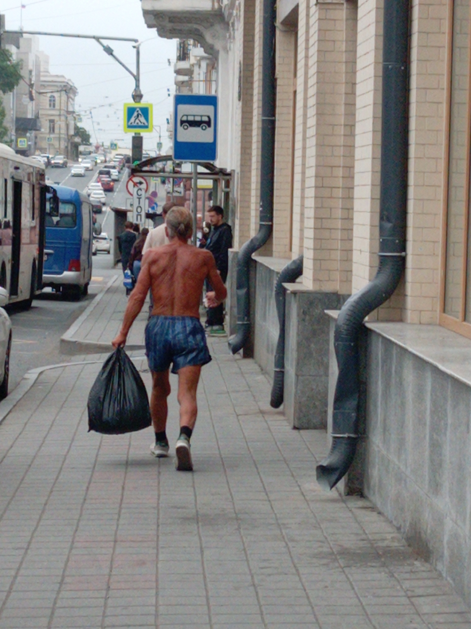 A man walks down a sidewalk in Vladivostok, Russia, carrying a large black trash bag in his right hand. He is shirtless and wearing blue patterned boxer shorts and dark socks with athletic shoes. The man is walking away from the camera and toward a street with traffic. A bus is parked to his left. The sidewalk is paved with rectangular gray stones. To the right of the sidewalk, there is a building with a light-colored brick facade, featuring downspouts and large windows. In the background, there are more buildings, traffic signs, and vehicles. The weather appears overcast.