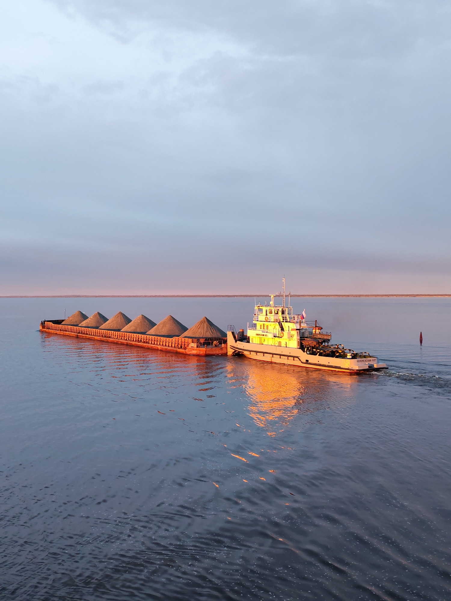 A long barge carrying what appears to be dark sand or gravel in conical piles is being pushed by a tugboat on a body of water, likely a river or large lake. The tugboat is white with multiple decks and structures. The water is mostly calm with ripples, and reflects the orange glow of the setting or rising sun on the tugboat. The sky is a gradient of light blue to pinkish-grey. A single red navigational buoy is visible in the distance to the right of the tugboat. No people are visible. The location and city of Bereg, Russia is not possible to determine based on the contents of the image.