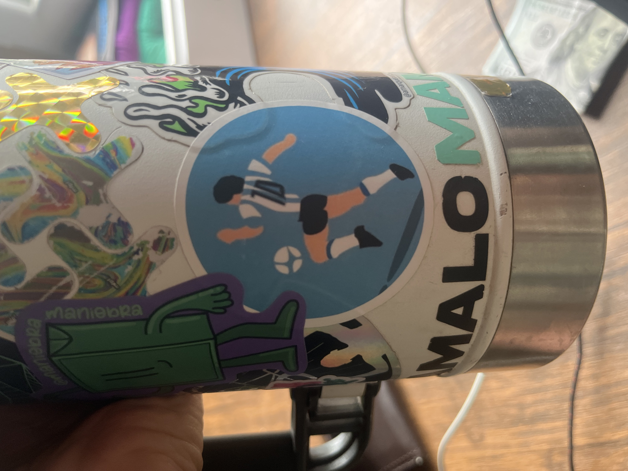 A close-up shows a cylindrical object covered in stickers, likely a water bottle or similar container. One prominent sticker depicts a soccer player in the blue and white stripes of Argentina's national team kicking a soccer ball. Another sticker shows a cartoonish green figure lying down. Adjacent to these is the word "MALOMA" written vertically in large, bold letters. The cylindrical object appears to be resting on a dark surface, possibly a table or desk. In the blurred background, a partially visible United States dollar bill is seen. There is no information in the image that indicates a location in Ushuaia, Argentina.