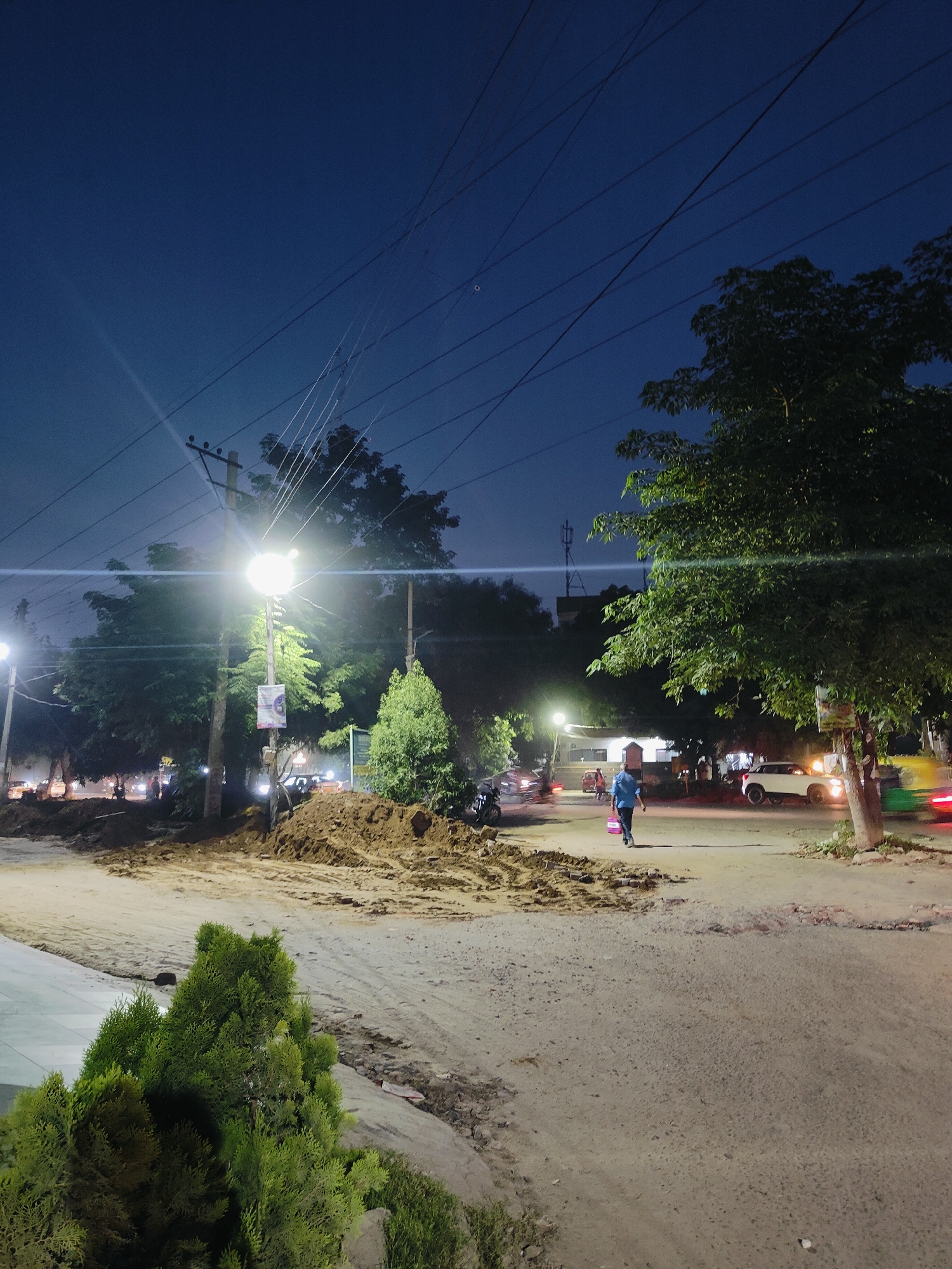 The image depicts a street scene at dusk or night in Gurgaon, India. A bright street light illuminates a pile of excavated earth in the foreground, suggesting ongoing construction or road work. A person in a blue shirt and dark pants is walking away from the camera on the right side of the road, carrying a pink bag. Several vehicles, including cars and a blurred auto-rickshaw or taxi, are visible on the street. Trees line the sides of the road, and power lines crisscross the dark sky. The overall impression is of a typical urban environment in India experiencing a nighttime transformation due to artificial lighting and activity.