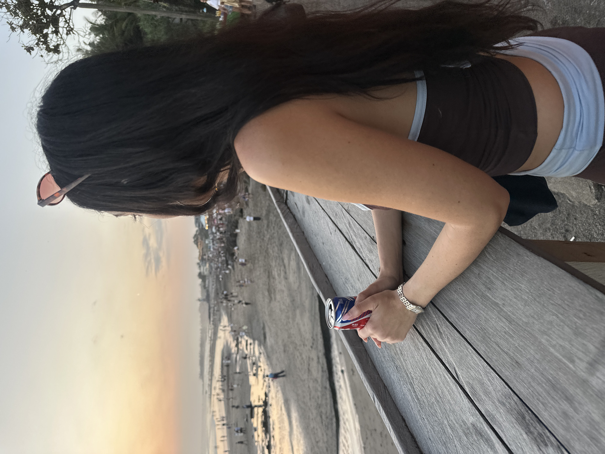 The image shows a woman from behind, sitting at a wooden table, with a beach and the ocean in the background. She has long, dark hair and is wearing sunglasses on top of her head. In her hands, she is holding a can of soda with a red, white, and blue design. Her arms are resting on the table. The beach is crowded with people, and the sun appears to be setting, casting a warm glow over the scene. The overall atmosphere is relaxed and casual.