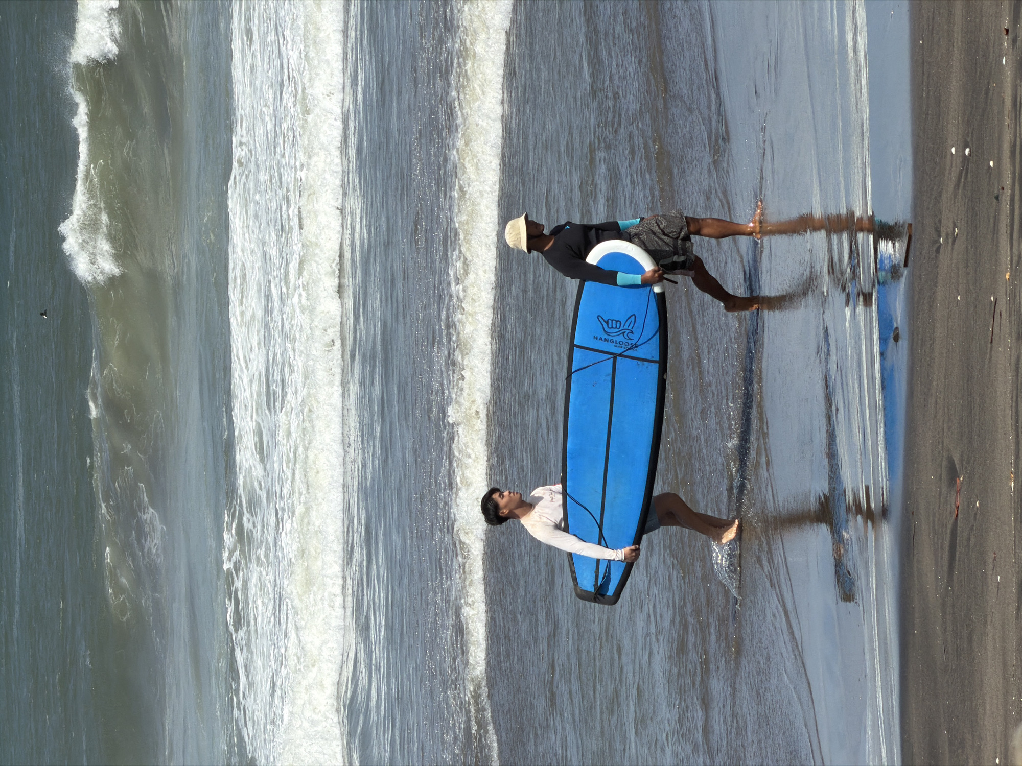 Two individuals are walking along the shoreline in the city of Mengwi, Indonesia, carrying a blue surfboard. The person on the left is wearing a long-sleeved white shirt and dark shorts. The person on the right is wearing a long-sleeved dark shirt, patterned shorts, and a light-colored bucket hat. They are walking from left to right across the frame, with the surfboards positioned between them. The ocean is in the background with moderate waves breaking. The sand in the foreground is dark and wet, with some small shells visible.