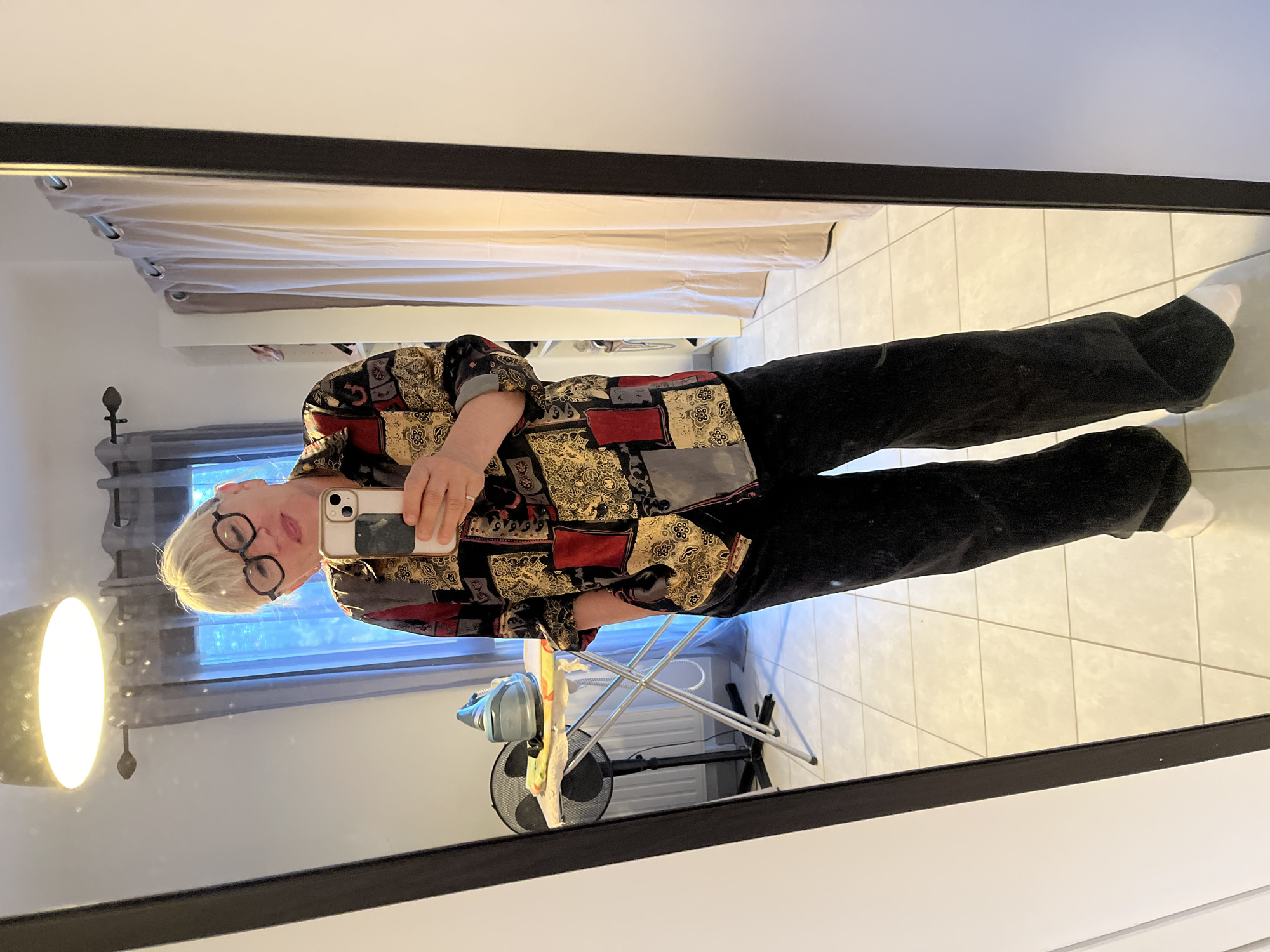 A woman in a patterned shirt and black pants is standing in a room and taking a mirror selfie. She is wearing glasses. The background includes a window with curtains, a floor lamp, a fan, an ironing board, a radiator, and tiled flooring. The room appears to be a utility or laundry room, given the presence of the ironing board and radiator. The location is likely Chambœuf, France, based on the user's query.