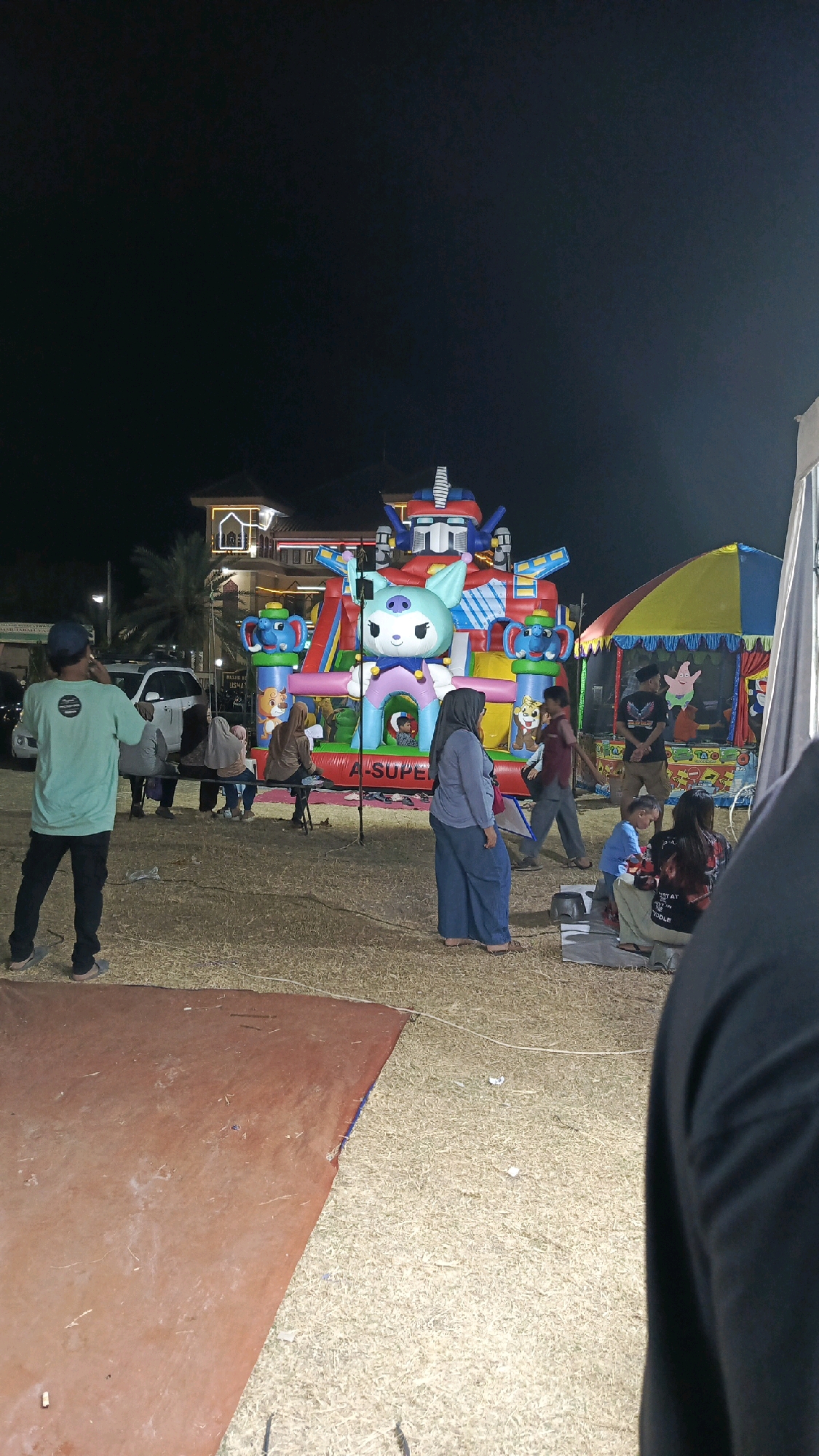 The image depicts an outdoor night scene in Tegal, Indonesia, likely at a local fair or event. The central focus is a large, brightly colored inflatable playground with a robot-like character at the top and a cute cat-like character at the entrance. Several people are gathered around the inflatable, with some children visible inside. To the right of the inflatable is a small tent with a yellow and red canopy, possibly a game stall or a performance area, with cartoon characters like SpongeBob SquarePants visible.
In the foreground, a man wearing a light green t-shirt and dark pants stands facing away from the camera, looking towards the event. Several other individuals are seated on benches or on the ground, observing the scene. A white SUV is parked behind the group of people to the left. The ground is covered in dry grass and a large, reddish-brown tarp or mat is in the lower left quadrant of the image. The background includes a building with architectural elements suggesting a mosque or a similar religious structure, illuminated by lights. The overall atmosphere suggests a lively community gathering under the night sky.