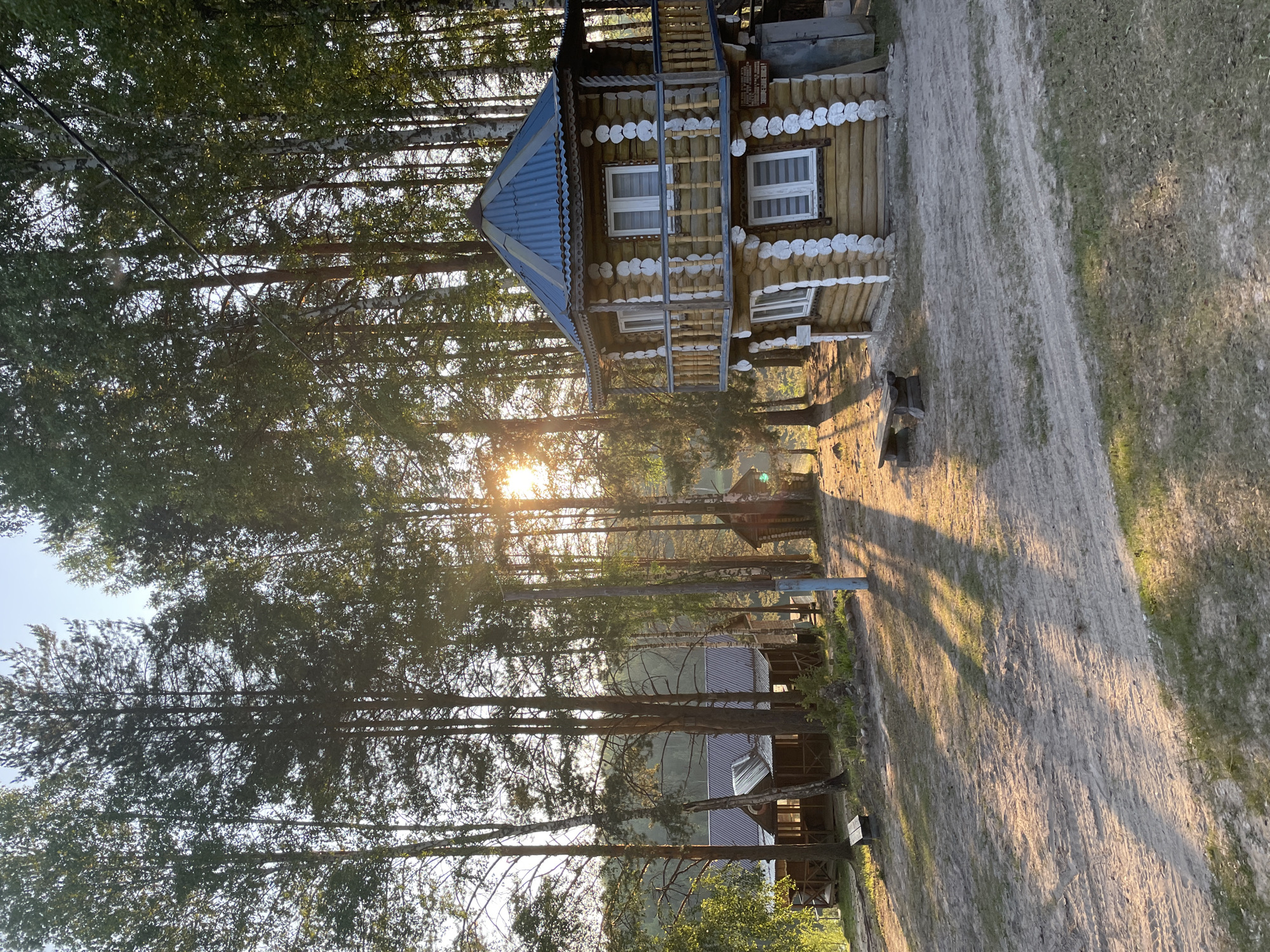 This is a medium shot, eye-level, outdoor, vertical photograph of a wooden cabin and surrounding forest in a rural area that may be the city of Пруды, Russia. The sun is low in the sky, casting long shadows across a dirt path and the front of the cabin. The cabin is a two-story structure made of logs, with white trim around the windows and a corrugated metal roof. There is a balcony on the second floor. To the left of the cabin, there is another, longer wooden building with a similar roof. The area is heavily wooded with tall pine and birch trees. The ground is covered in dirt and sparse grass. There are no people visible in the image.