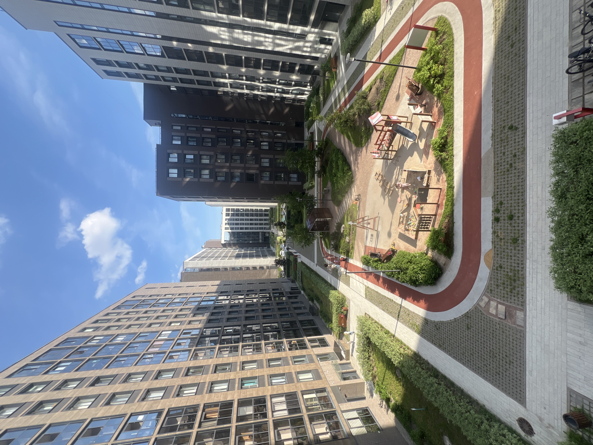 This is an overhead forensic view of a modern residential courtyard in Saint Petersburg, Russia, observed during daytime under a clear blue sky with scattered white cumulus clouds.
The scene is framed by multi-story apartment buildings of contemporary design. The prominent building on the left features a light brown or tan facade with dark grey window frames and numerous glass-fronted balconies, presenting a uniform grid. The building on the right exhibits a contrasting light grey or white facade with vertical dark banding and dark window frames. In the background, additional modern residential structures with varying dark brown, grey, and light facades complete the urban residential complex.
The central courtyard is meticulously landscaped, featuring a network of pedestrian pathways and green spaces. Light grey concrete pavers form the main walkways, some incorporating textured grey permeable pavers with sparse grass growth. A distinct, reddish-brown running or cycling track with white borders curves through the courtyard, separating paved areas from planted sections.
At the center of the courtyard is a children's playground with a large sandpit. Within the playground, wooden play structures are visible, including a slide, a climbing frame, and what appears to be part of a swing set. Several individuals are present: approximately six to eight people, including adults and young children. Two children are actively playing in the sandpit, and another child is near the slide, with an adult supervising nearby. Other adults are seen walking along the paved pathways.
Throughout the courtyard, modern black lampposts are strategically placed. Wooden benches offer seating along the paths and in grassy areas. The landscaping consists of neatly trimmed hedges, small bushes, and young trees. In the bottom-left corner, a wooden slatted waste bin and a manhole cover are visible on a paved path. In the bottom-right corner, a bicycle rack holds at least two bicycles.
The sun casts distinct shadows, indicating a bright, sunny day. The overall impression is one of a newly developed, well-maintained, and family-friendly urban environment.