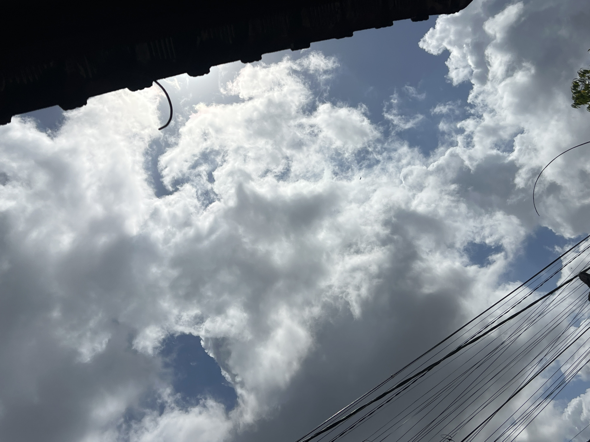 The image, taken in Denpasar, Indonesia, shows a dramatic sky filled with a mix of dark, heavy clouds and bright, sunlit cumulus formations. The sun is positioned behind the clouds on the right side of the frame, casting a bright glow and creating a halo effect. In the foreground, a series of black power lines are strung diagonally across the lower left portion of the image, creating a stark contrast with the natural elements above. To the far right, a dark, silhouetted structure, possibly a building or rooftop, juts into the frame from the top edge, its details obscured by shadow. A thin, dark, curved wire or rod extends from this structure into the sky. The overall impression is one of an overcast yet luminous day with urban infrastructure intruding upon the natural grandeur of the sky.