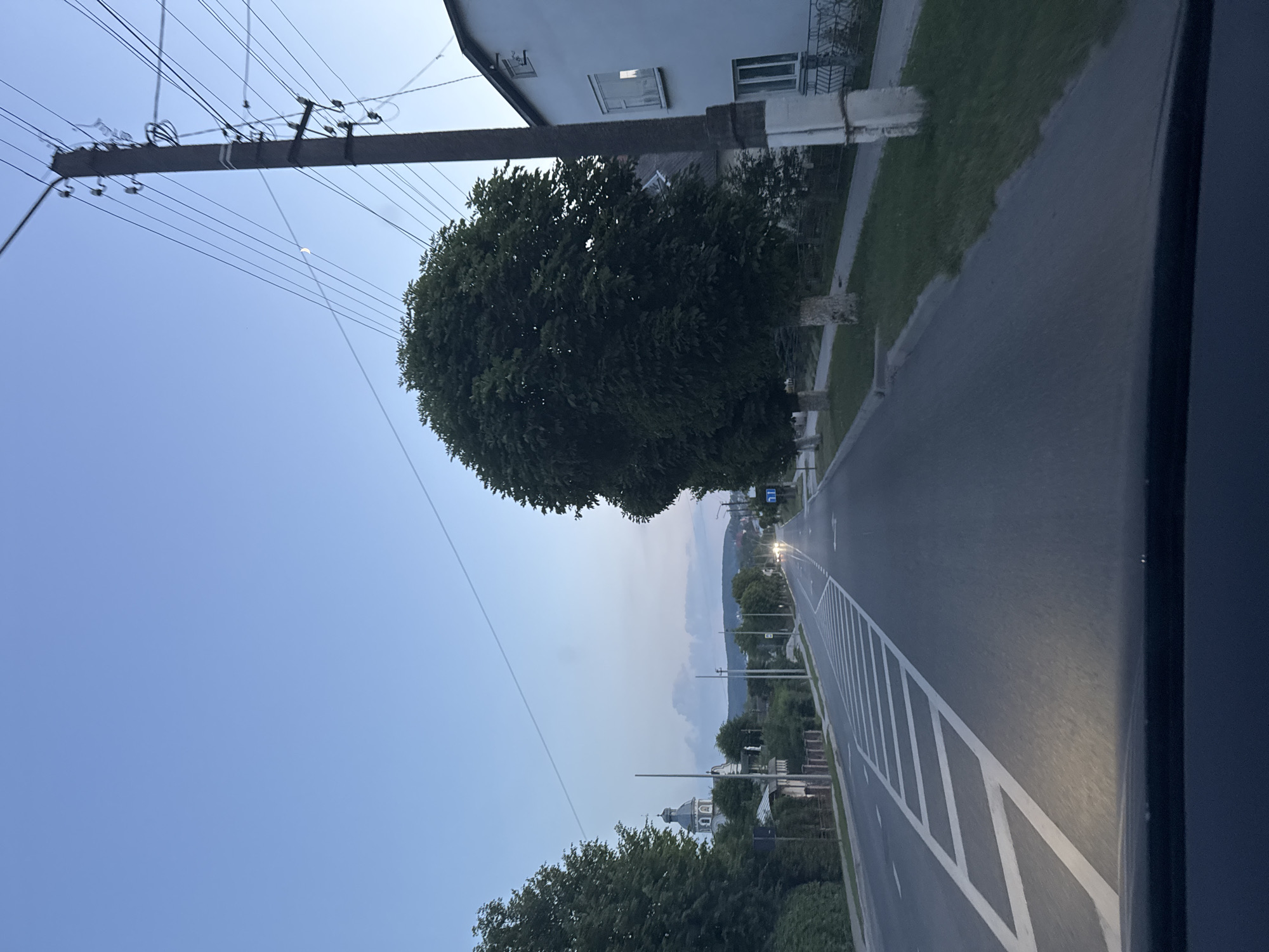 The image depicts a view from a vehicle traveling along a road in the city of Berezhany, Ukraine. The time of day appears to be dusk or early evening, with a clear, pale blue sky.
On the left side of the road, there are trees and buildings, including what appears to be a church with a domed roof and a tall spire. Further down the road, other buildings and more trees are visible.
On the right side of the road, there is a white building with windows and a large, dark green tree. A utility pole with multiple wires runs along the side of the road.
The road itself has white lane markings, including a pedestrian crossing marked with diagonal white lines. The road curves gently to the right as it recedes into the distance. There are streetlights and vehicle headlights visible in the distance, indicating ongoing traffic. The lower part of the image shows the dashboard or hood of the vehicle from which the photo is taken. There are no people visible in the image.