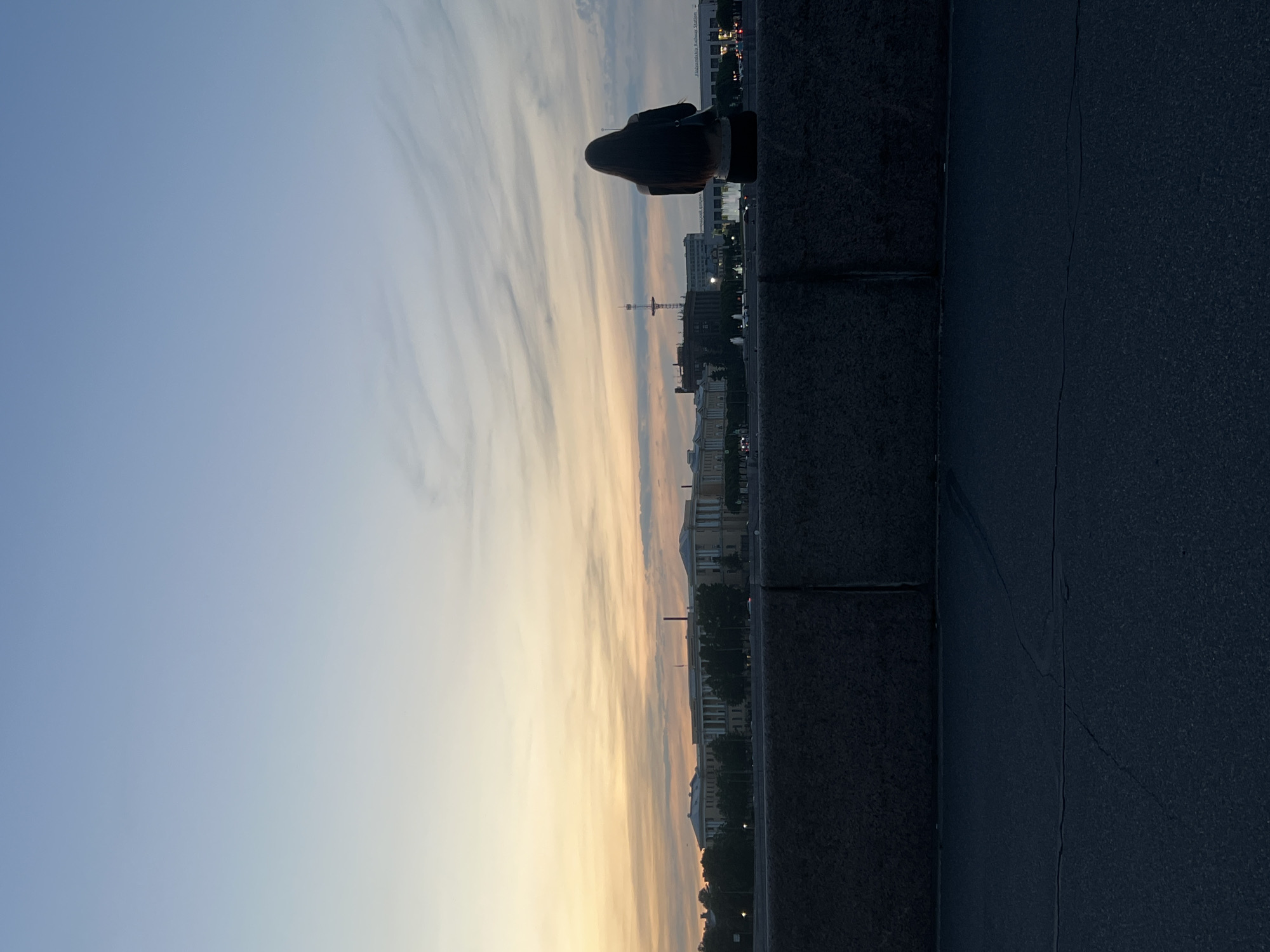 A person is seated on a stone embankment, facing away from the camera, looking out at a cityscape during twilight. The sky is a gradient of light blue and soft orange, with wispy clouds. The cityscape includes buildings with classical architecture, a tall antenna, and lights from streetlamps and possibly vehicles. The embankment is made of dark, rough-hewn stone blocks. In the foreground, a dark asphalt surface with visible cracks is present. The overall atmosphere is calm and serene. The location is identified as being in the city of Unknown, Russia.