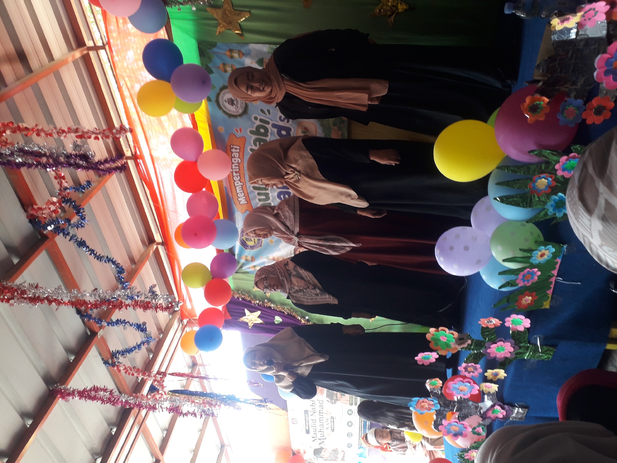 A group of women wearing hijabs are on a stage, likely in Cankuang, Indonesia, for an event. The stage is decorated with colorful balloons and tinsel. A banner behind the women reads "Memperingati Maulid Nabi Muhammad SAW," indicating the event is a celebration of the Prophet Muhammad's birthday. One woman is holding a microphone, suggesting she is speaking or singing. Other people, including children, are visible in the background. The setting appears to be an indoor space with a tin roof and wooden beams.