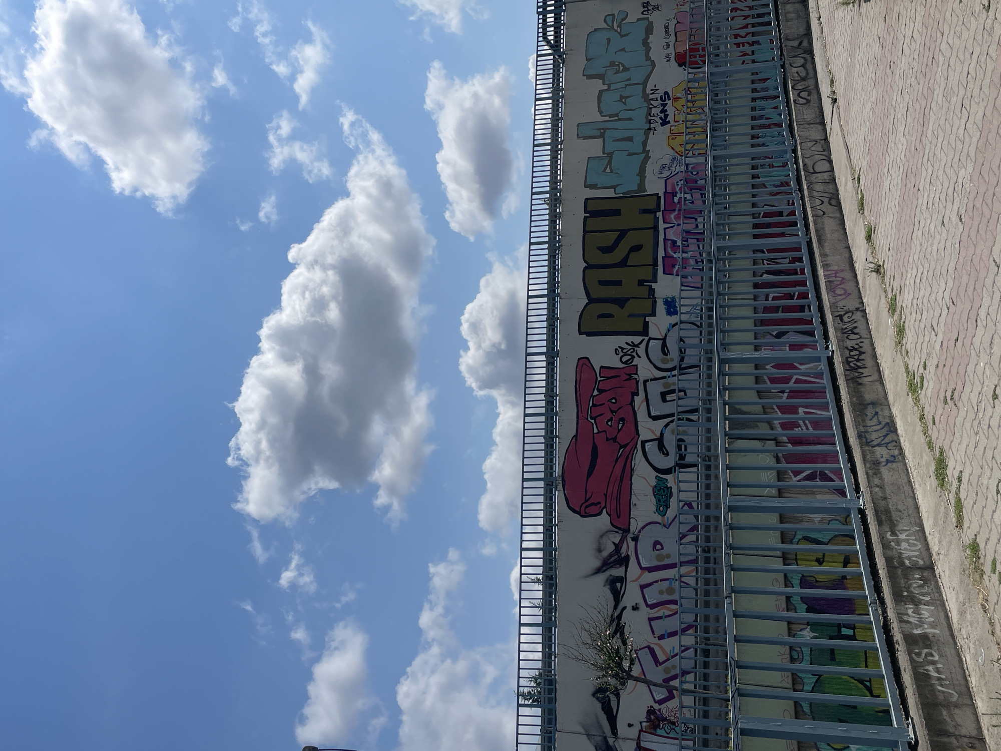 The image shows a section of a graffiti-covered wall under a bright blue sky with scattered white clouds. A blue metal railing runs horizontally across the frame, separating the viewer from the wall, which appears to be part of an overpass or bridge. The wall itself is extensively decorated with various graffiti tags and pieces in different colors and styles. One prominent piece in the upper section features large, stylized lettering in yellow and teal, reading "RASH GALAXY". To the left of this, there is a large red graphic that resembles a cartoonish head or mask. Below this, the wall is covered with more graffiti in pink, purple, and white. Another railing runs lower down, in front of another layer of graffiti, this one predominantly in shades of green and pink. The ground in the foreground is paved with reddish-brown interlocking bricks, with some patches of dry grass and concrete. The overall scene depicts an urban environment with a focus on street art. The context of Kadıköy, Turkey, suggests this is likely a public space within the city.