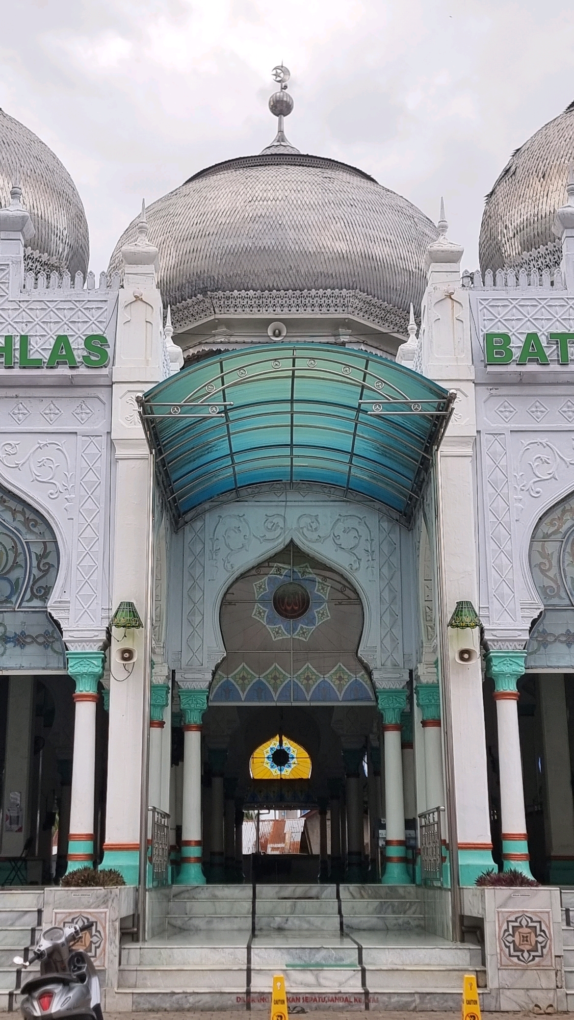 The image shows the exterior of the Baiturrahman Grand Mosque located in Lhokseumawe, Indonesia. The mosque features a central dome with a crescent moon and star finial, flanked by two smaller domes. The facade is white with intricate decorative patterns. A prominent arched entryway is covered by a turquoise-colored retractable awning supported by metal beams. Arabic calligraphy is visible above the arched entrance. The mosque has tall, fluted white columns with colored bands at the top and bottom. Steps lead up to the entrance. A silver scooter is parked on the left side of the steps, and yellow caution signs are placed near the bottom of the stairs. The sky is overcast.